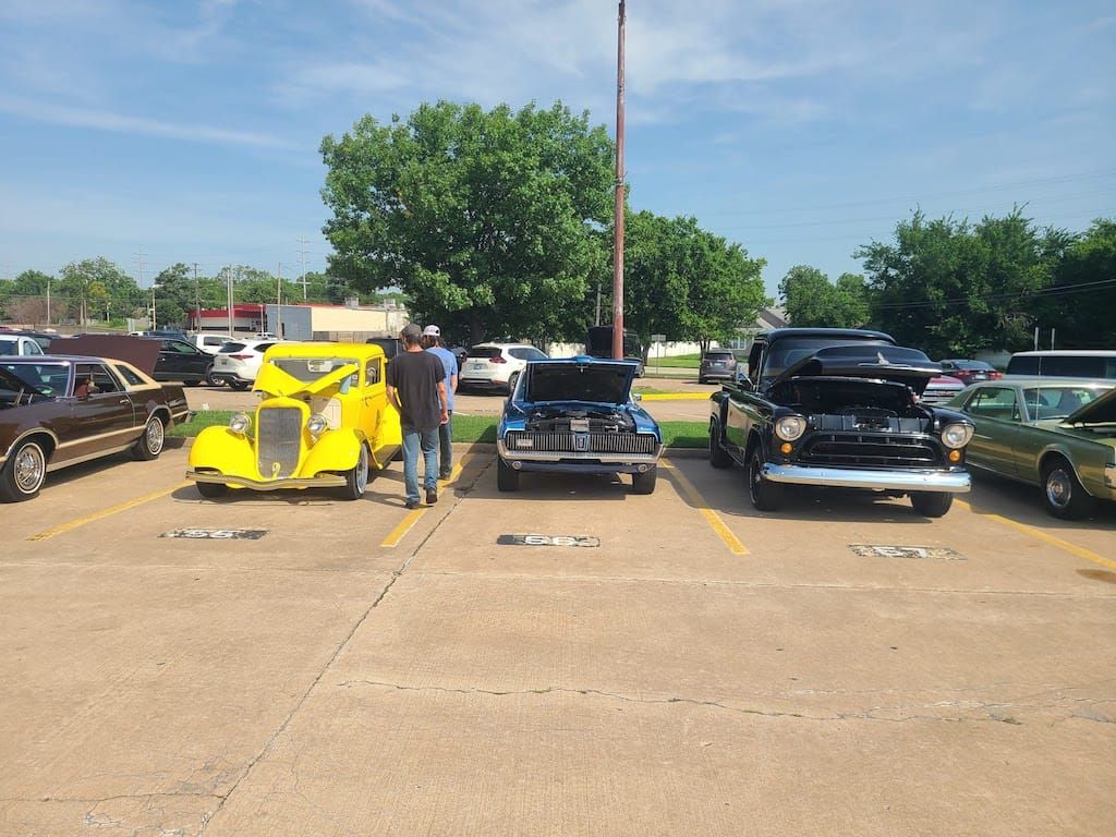 Classic cars parked in a lot; two men inspect the hoods of vehicles, bright yellow car in front.