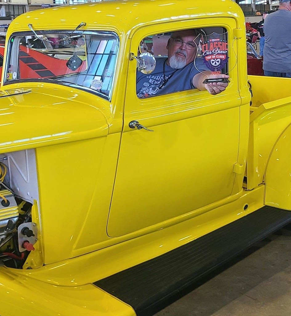 Man in yellow classic pickup truck at an auto show, holding something.