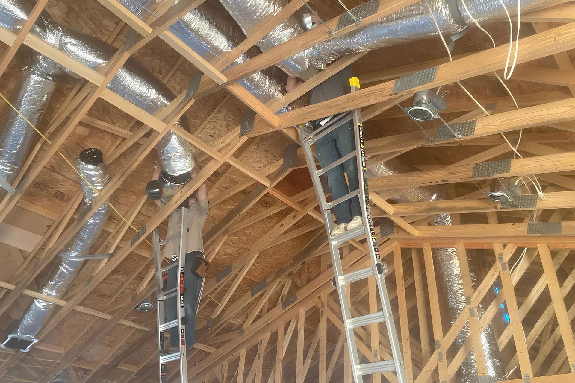 Workers installing HVAC ductwork in a wood-framed ceiling. They are using ladders in a construction setting.