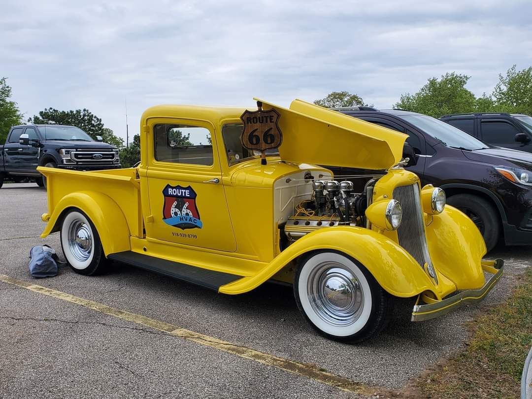 Yellow vintage Route 66 pickup truck with open hood, white-walled tires, parked outdoors.