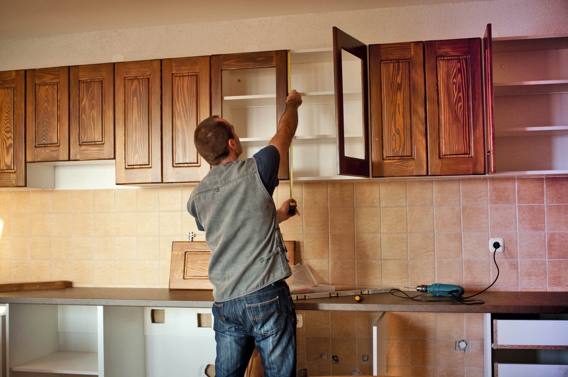 Man measuring a kitchen cabinet during a remodel; cabinets, countertop, and tools visible.