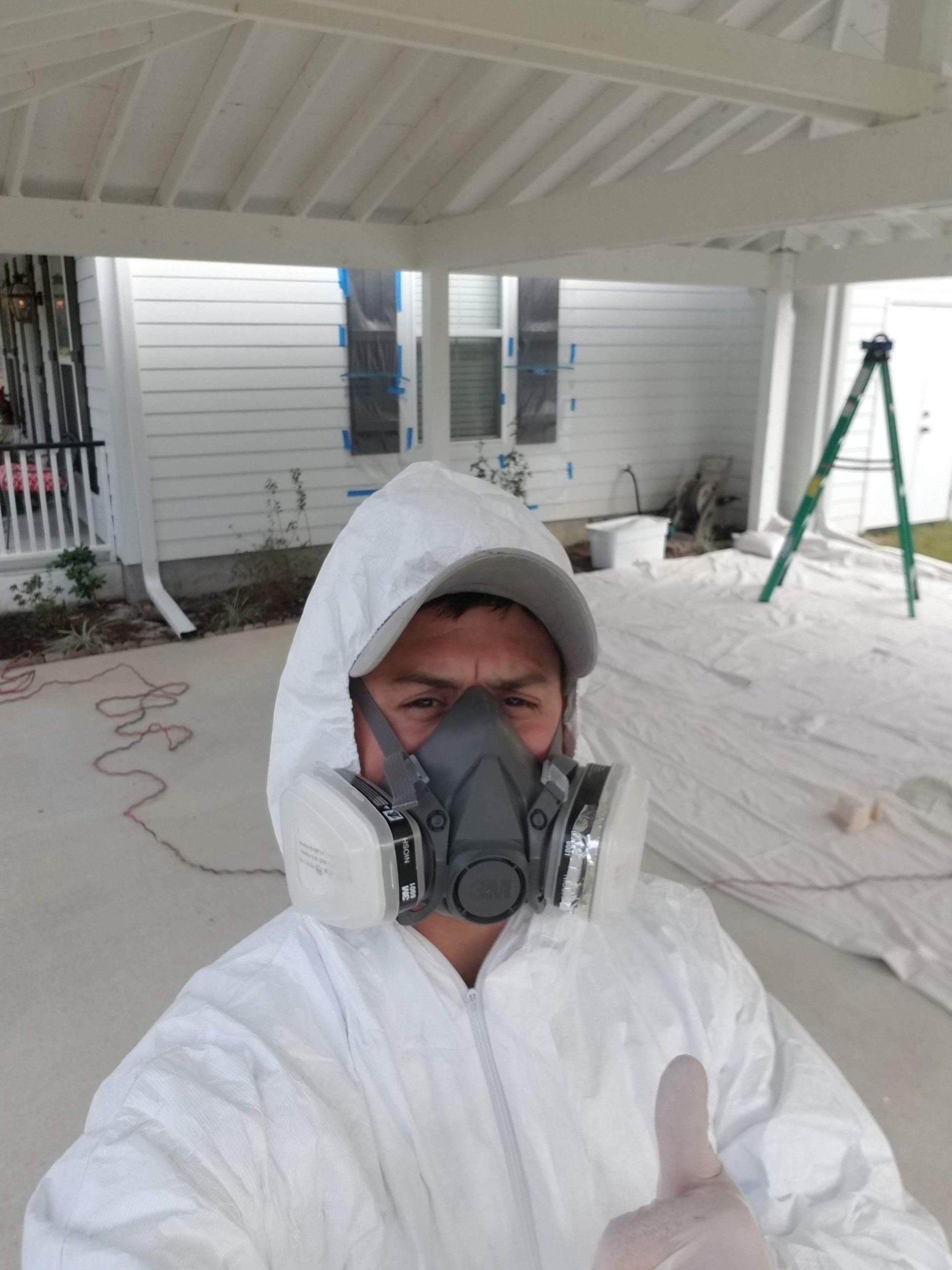 Person in a white protective suit with a respirator giving a thumbs-up gesture outdoors; painting a white structure.