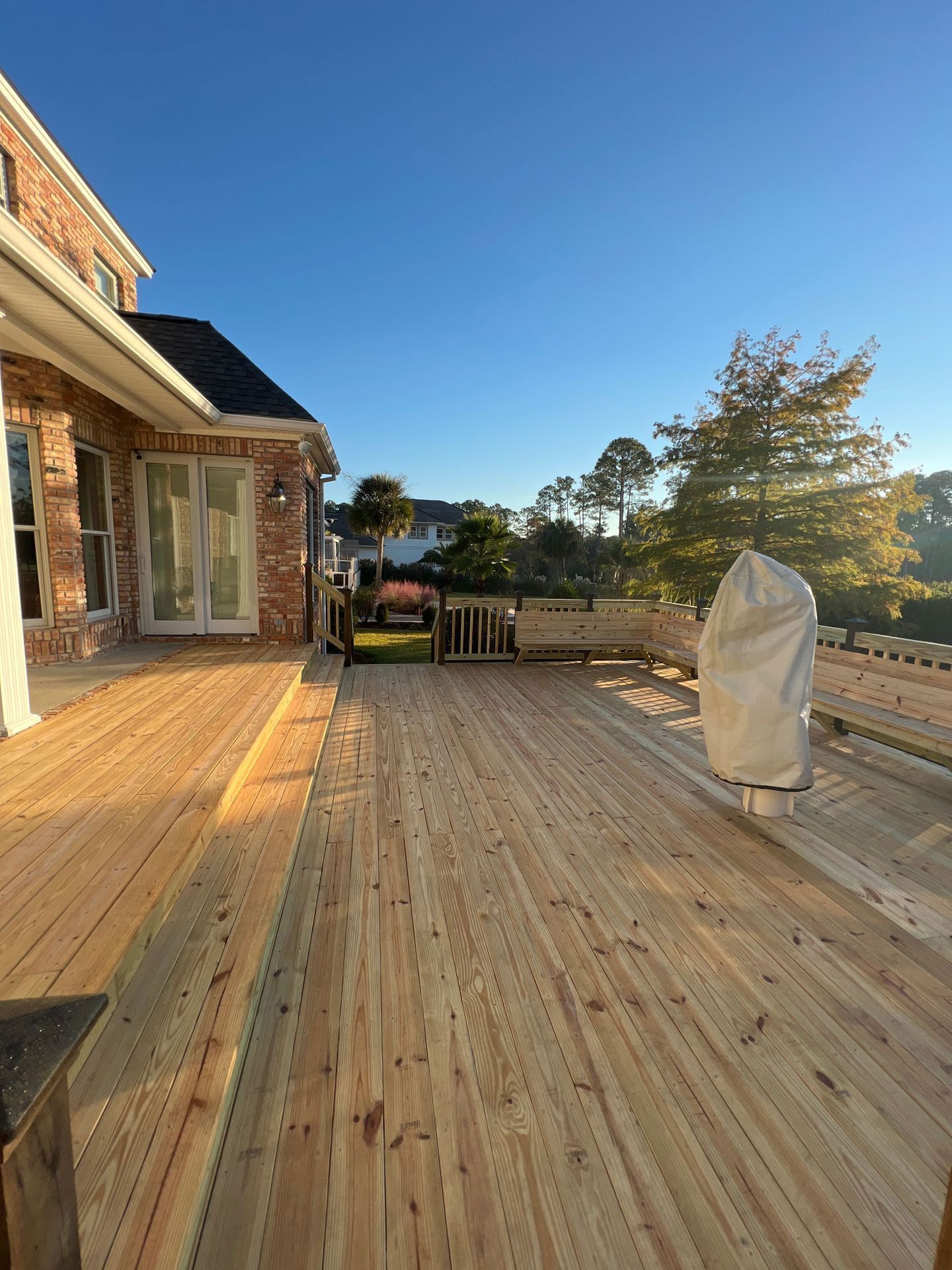 Wooden deck extending from a brick house, overlooking a yard and trees under a clear, blue sky.
