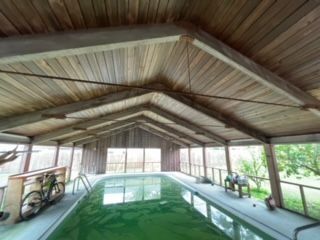 Indoor swimming pool under wooden roof; green water, bike, and outdoor scenery visible.