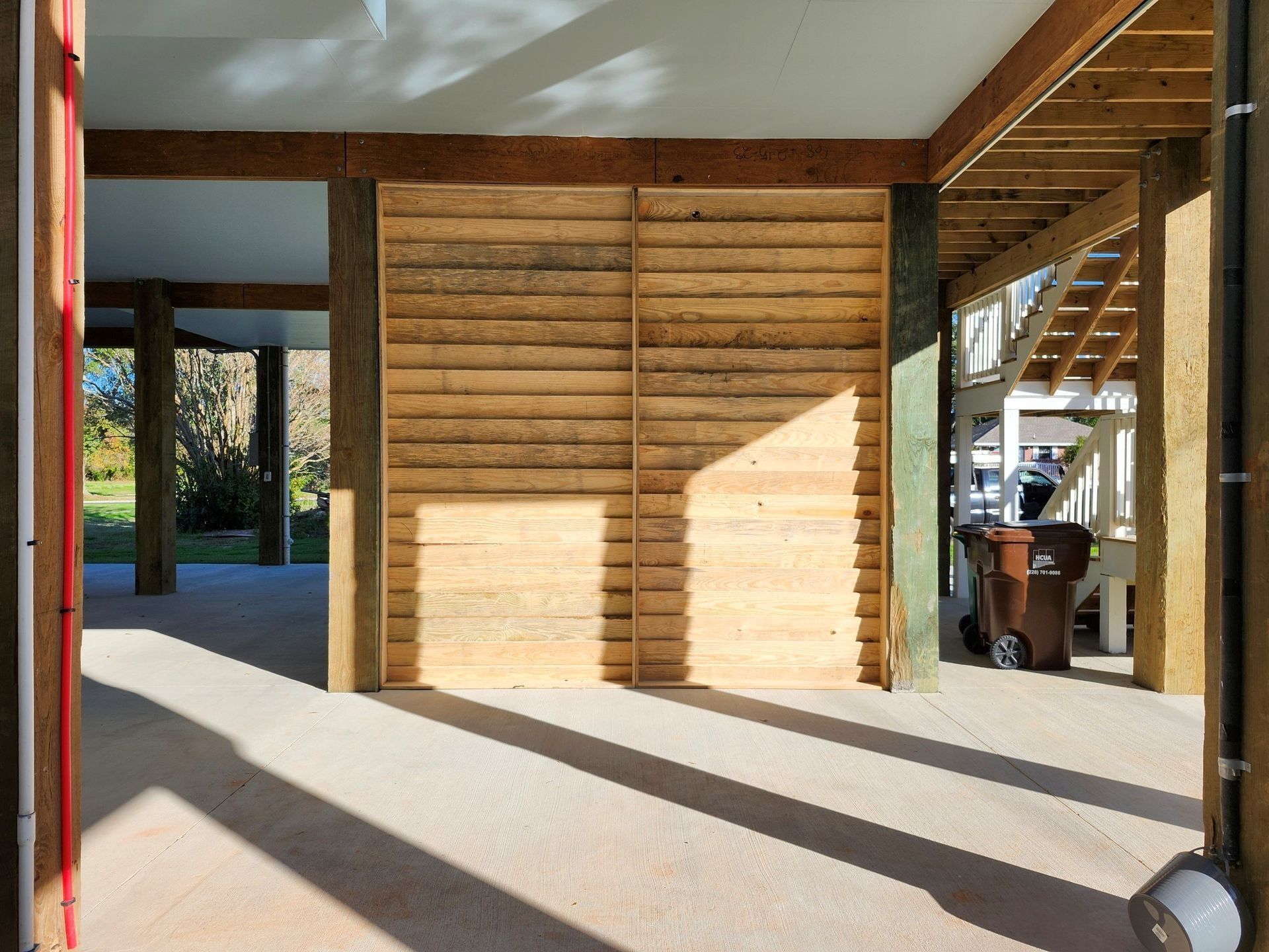 Open-air structure with wooden slat doors, concrete floor, sunlight casting shadows. A trash can is visible.