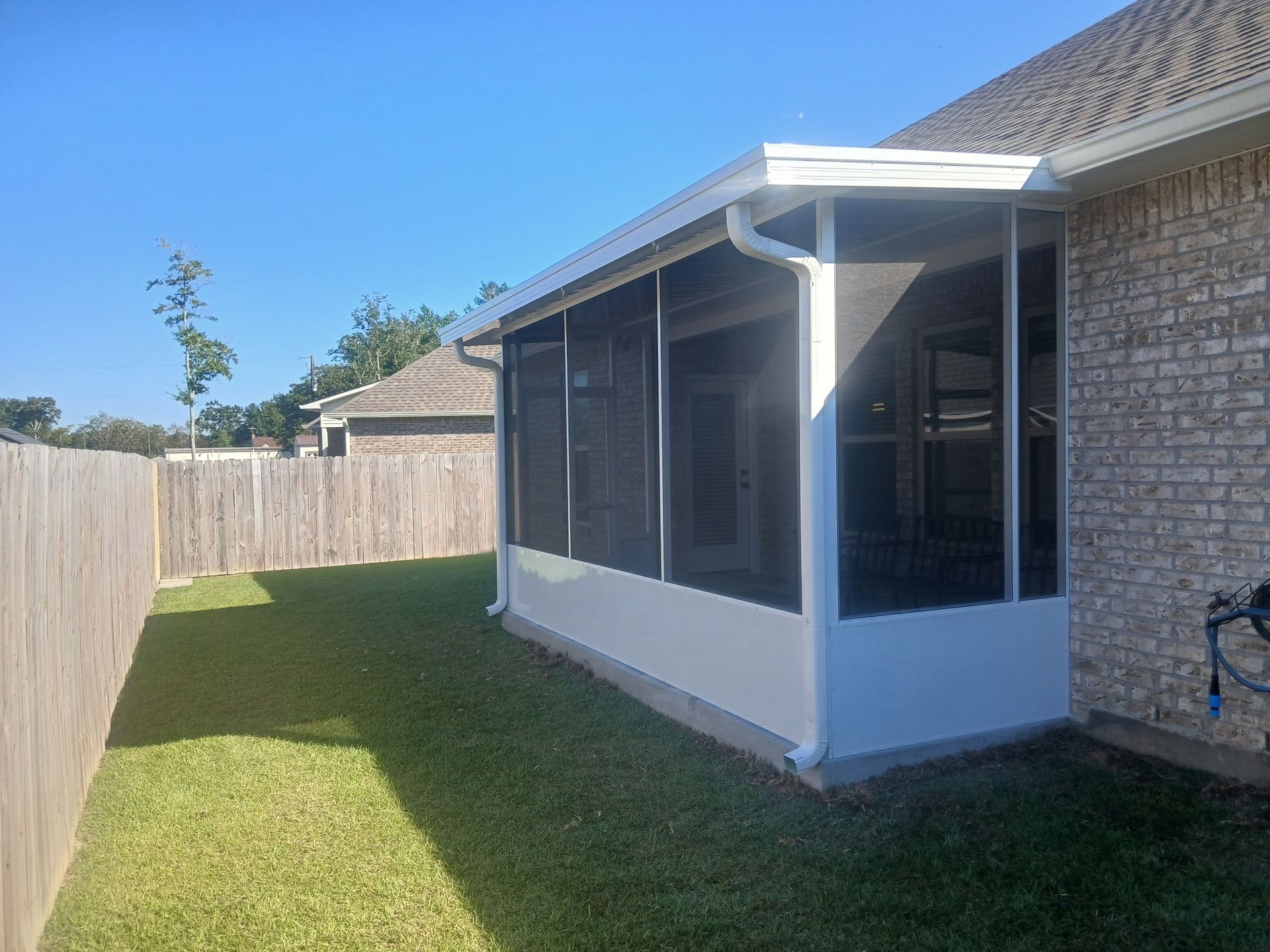 Screened-in porch attached to a brick house in a backyard with green grass and a wooden fence.