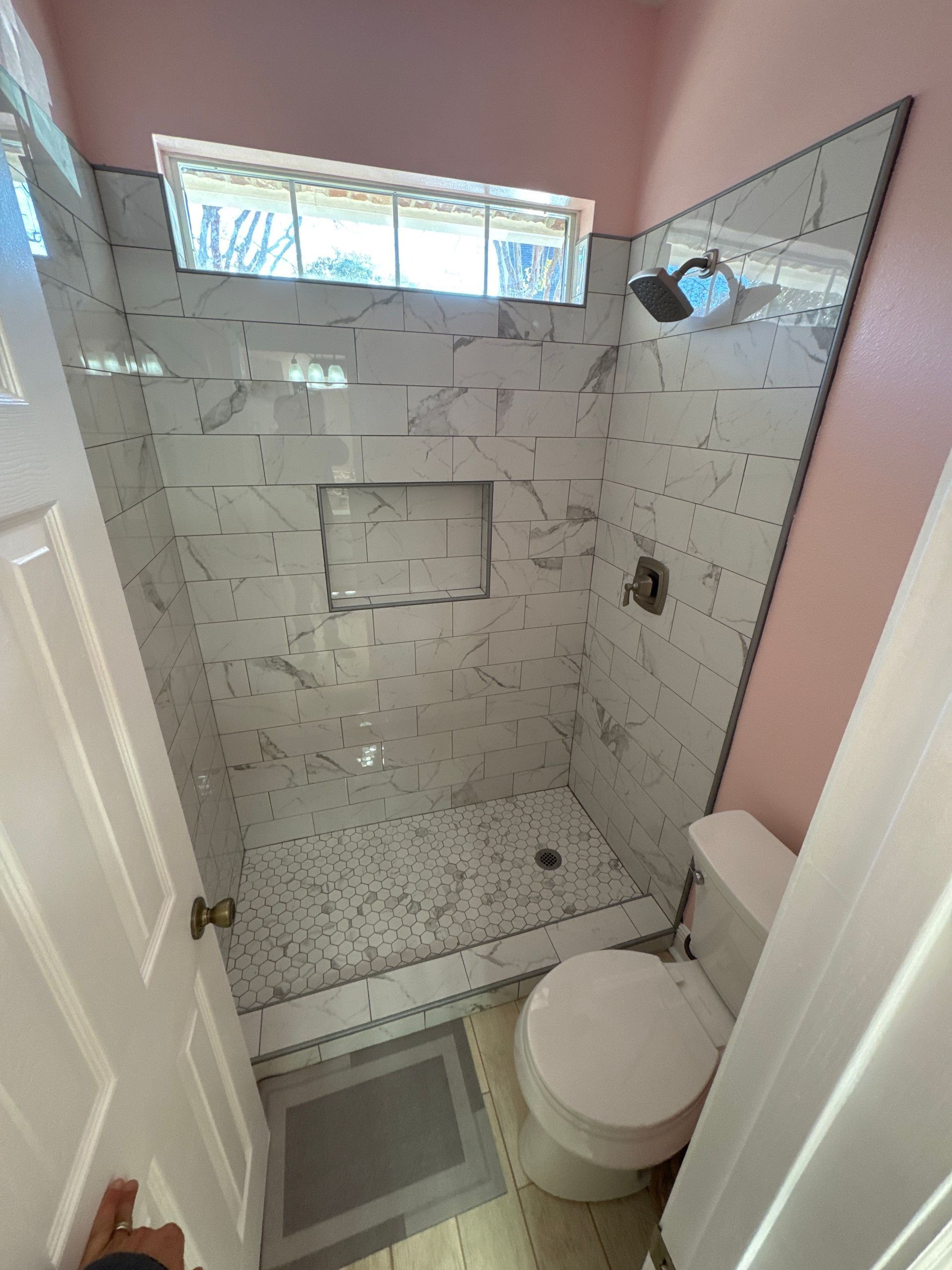 Small bathroom with marble-tiled shower and toilet, pink walls, and a small window.