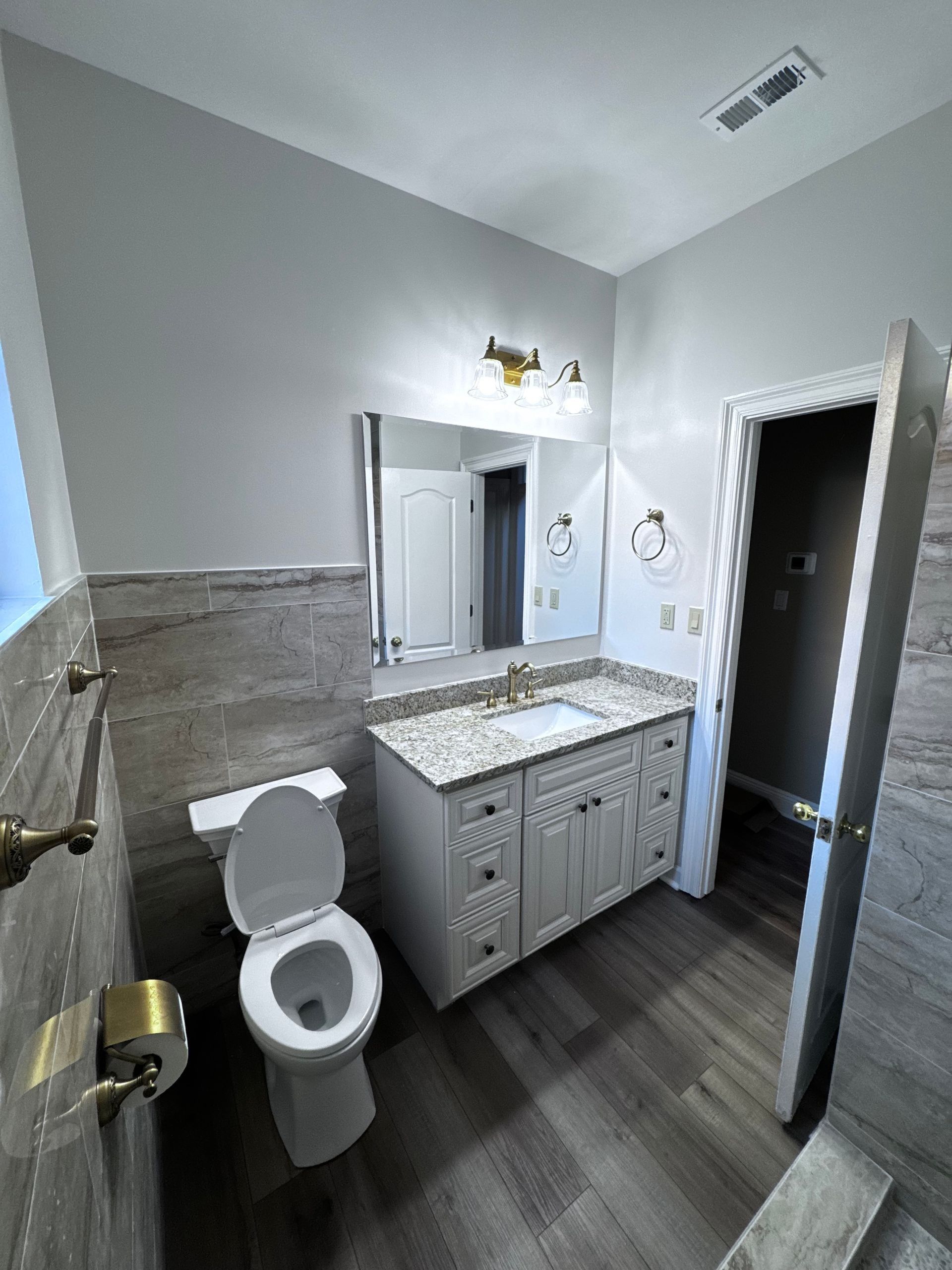 Bathroom with gray walls, wood-look floor, white vanity with granite countertop, toilet, and open door.