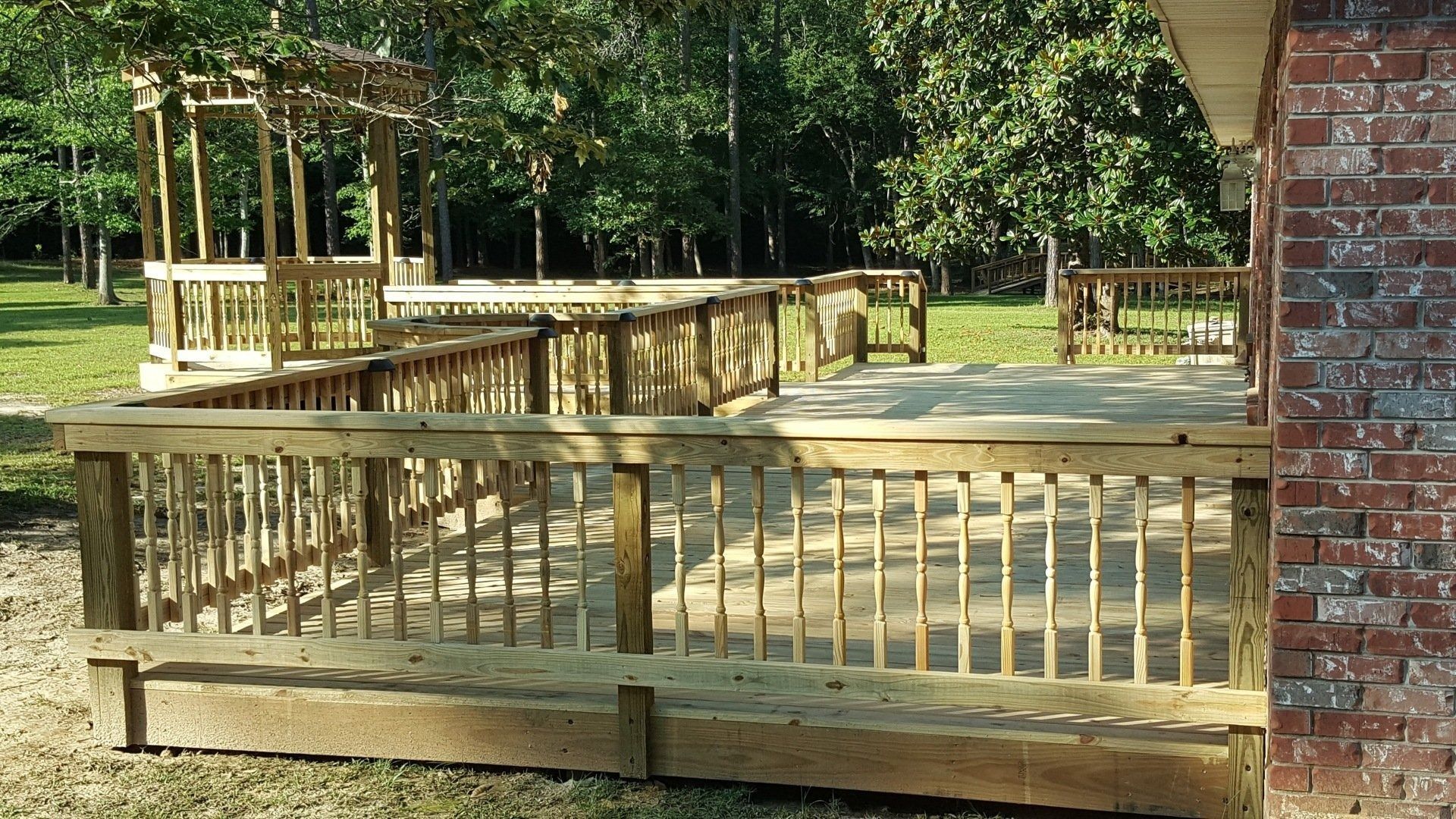 Wooden deck with railings and a pergola, attached to a brick building, surrounded by green grass and trees.