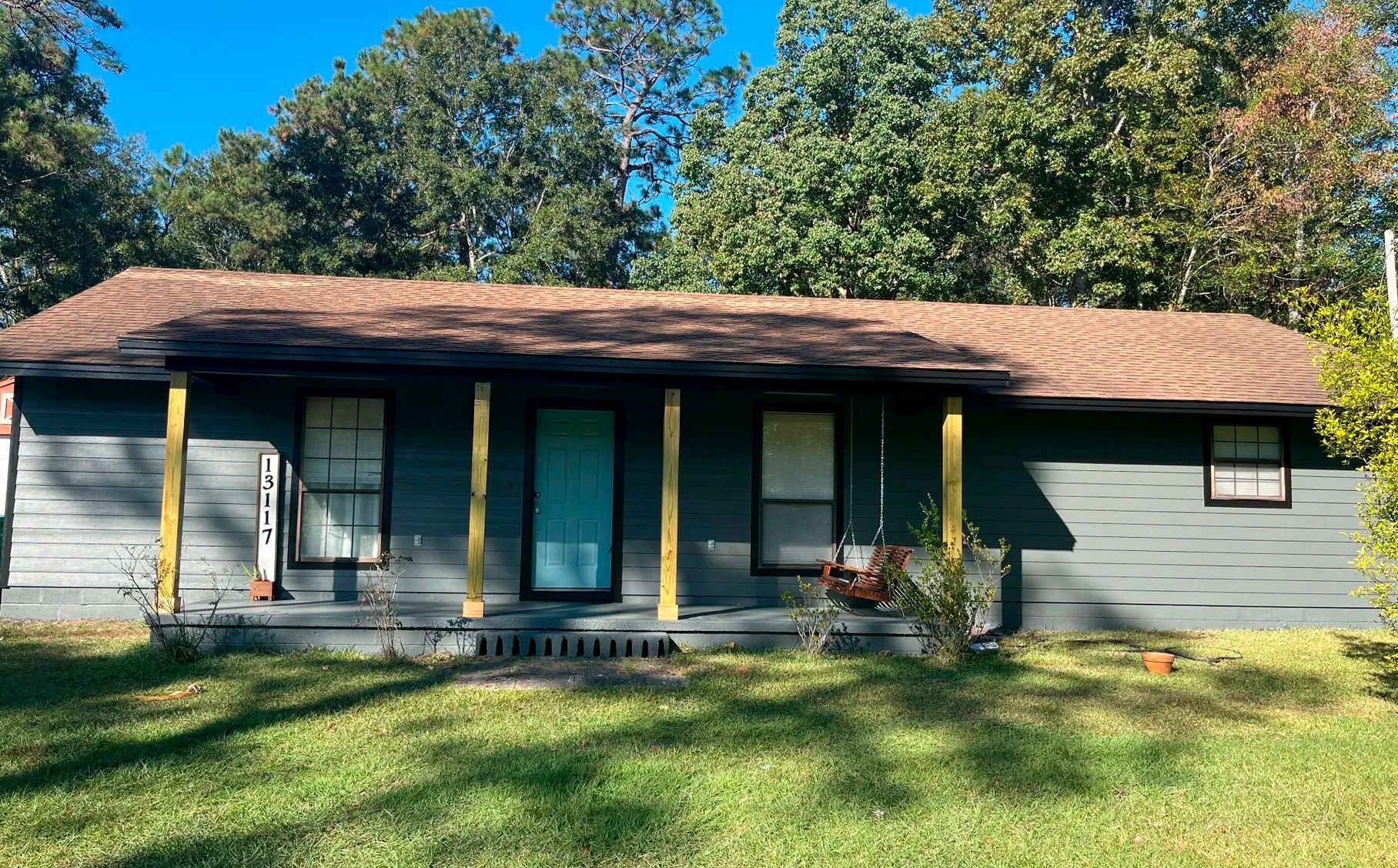 A one-story house with a porch and green siding, set against a backdrop of trees.