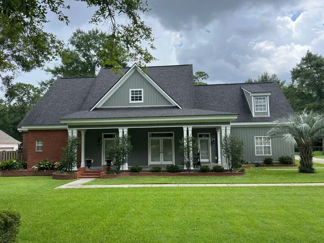 Green house with brick accents, white trim, and a porch, set on a green lawn with trees.