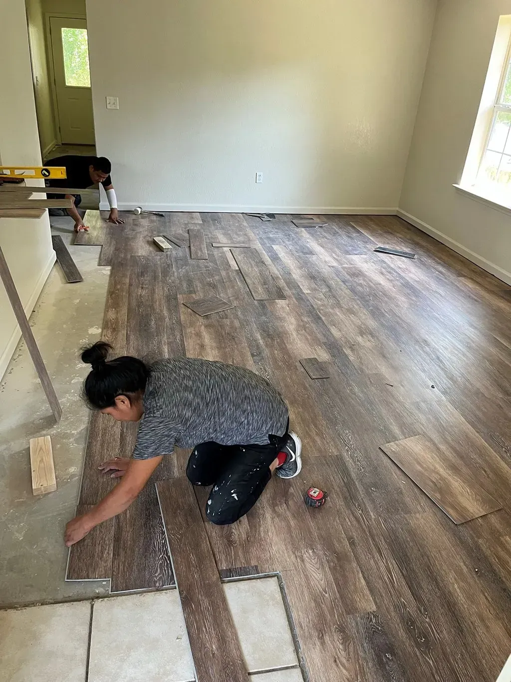 Woman installing wood-look flooring in a room. Another person is in the background. Light-colored walls, window on the right.
