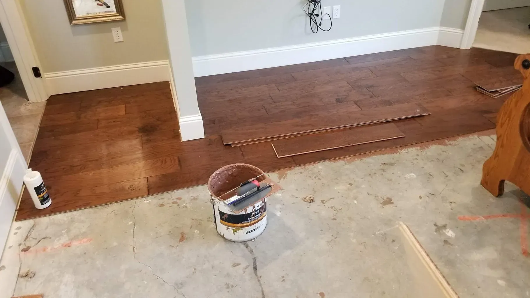 Hardwood floor installation in progress, showing planks, adhesive bucket, and concrete subfloor.