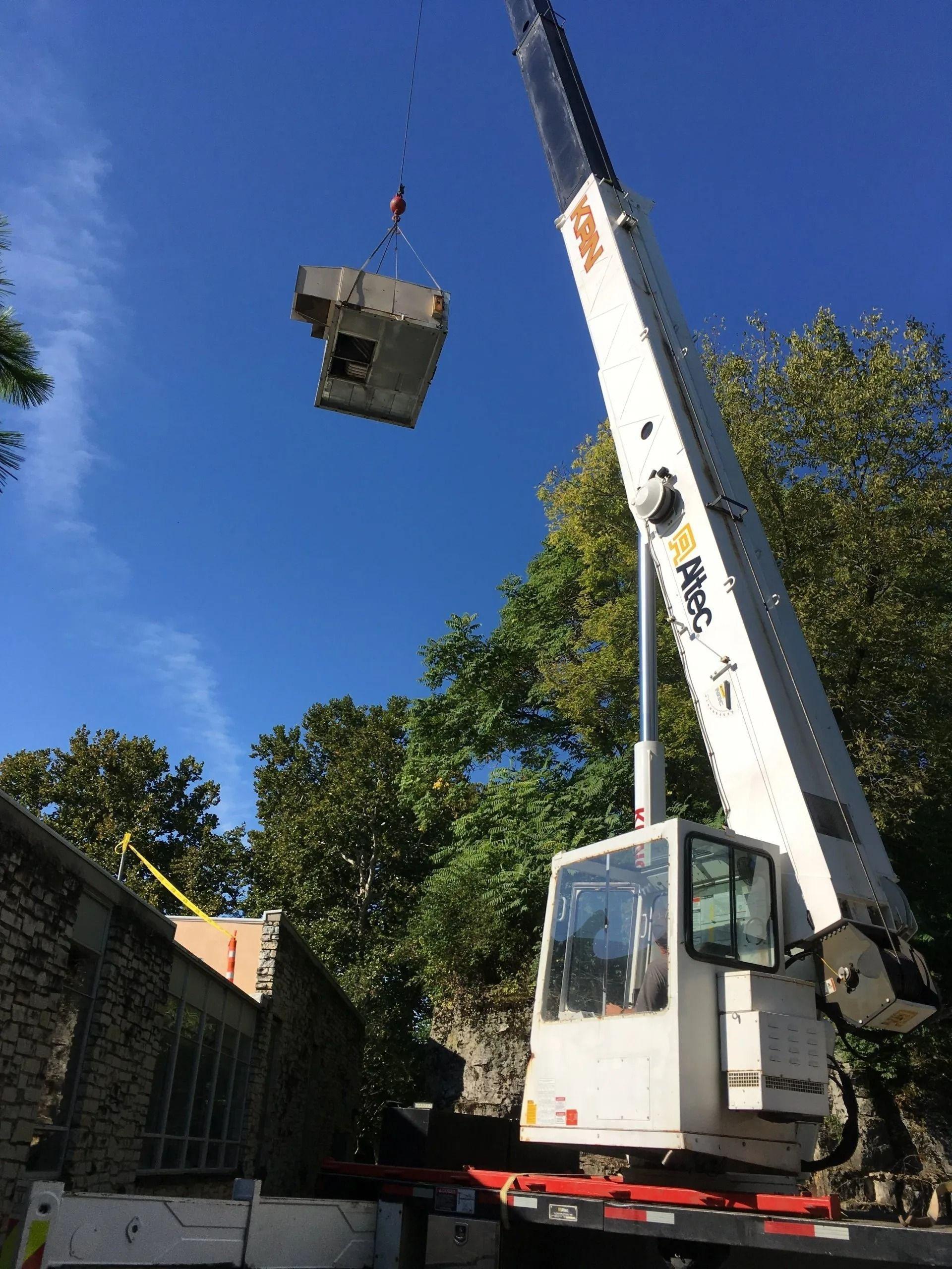 A crane lifting a large, square concrete structure above a brick building, under a blue sky.