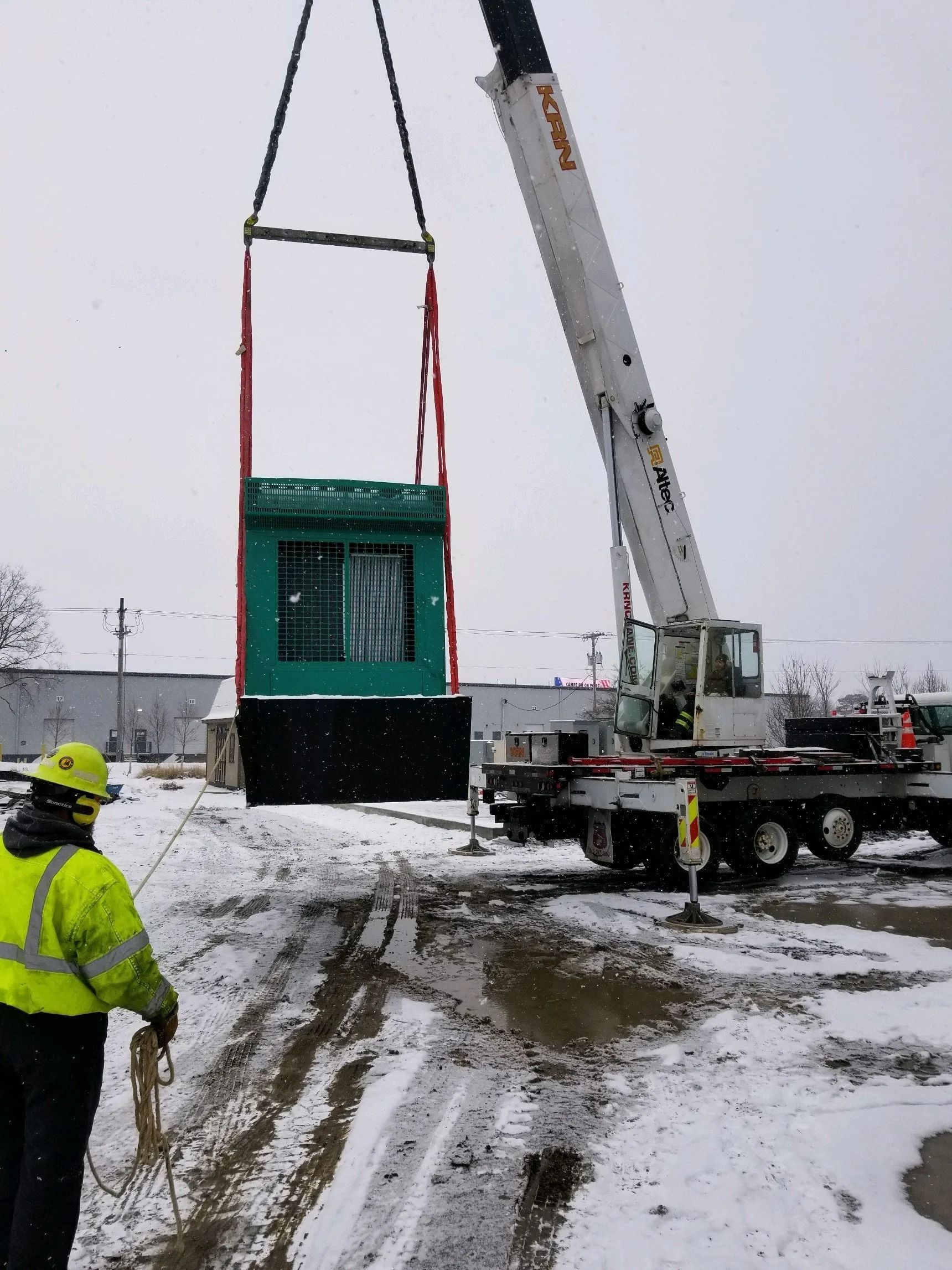 Crane lifting a green booth structure, with a worker in a safety vest. Snow on the ground.