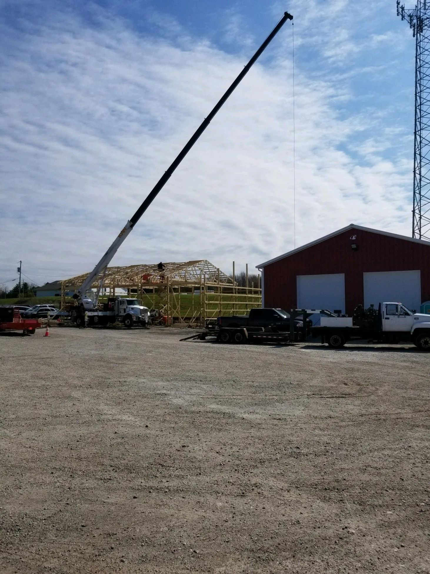 Crane lifting materials at a construction site near a red building and gravel parking lot.