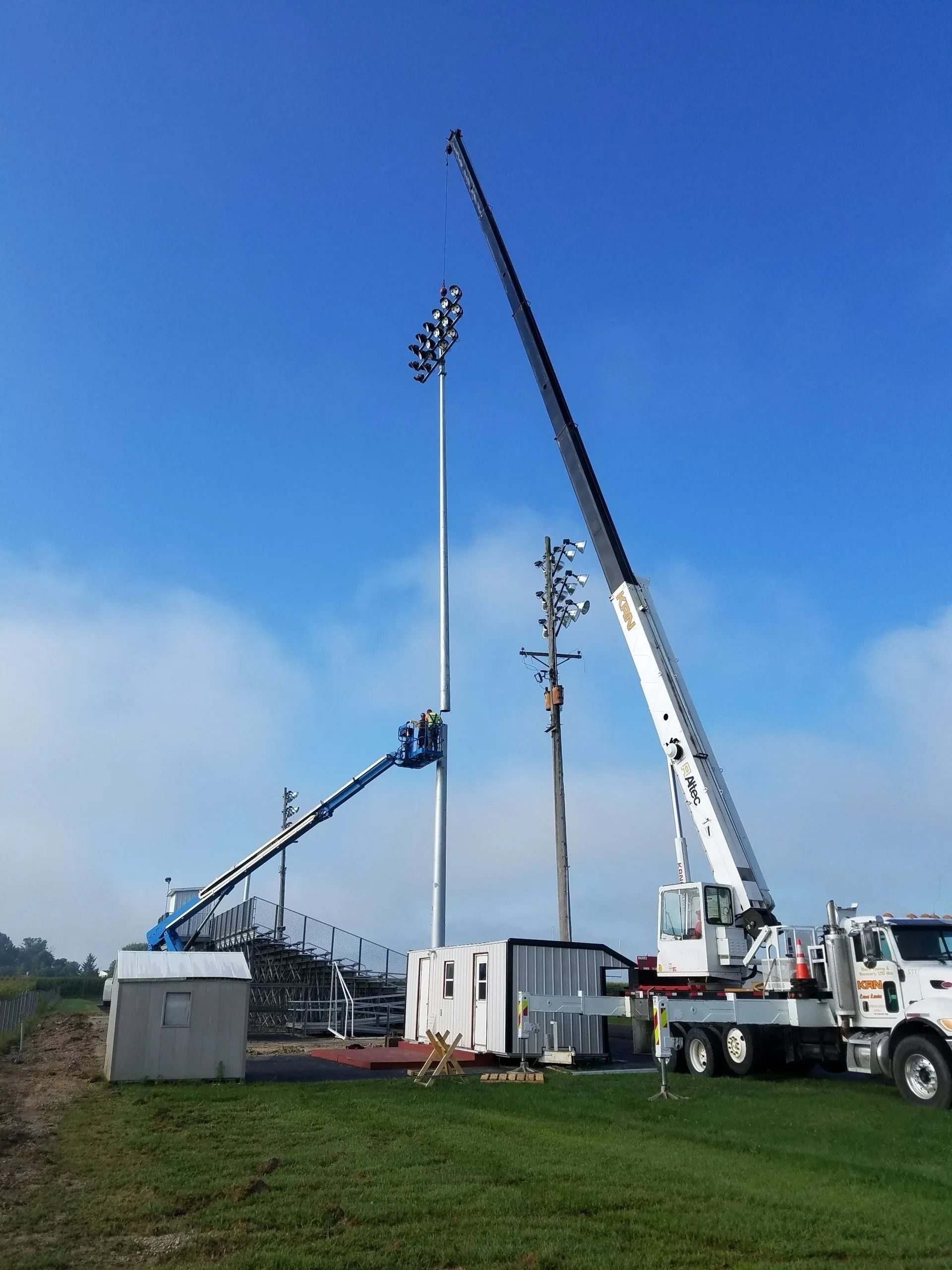 Construction site: crane and lift trucks around a tall light pole, utility pole, and a small building under a blue sky.