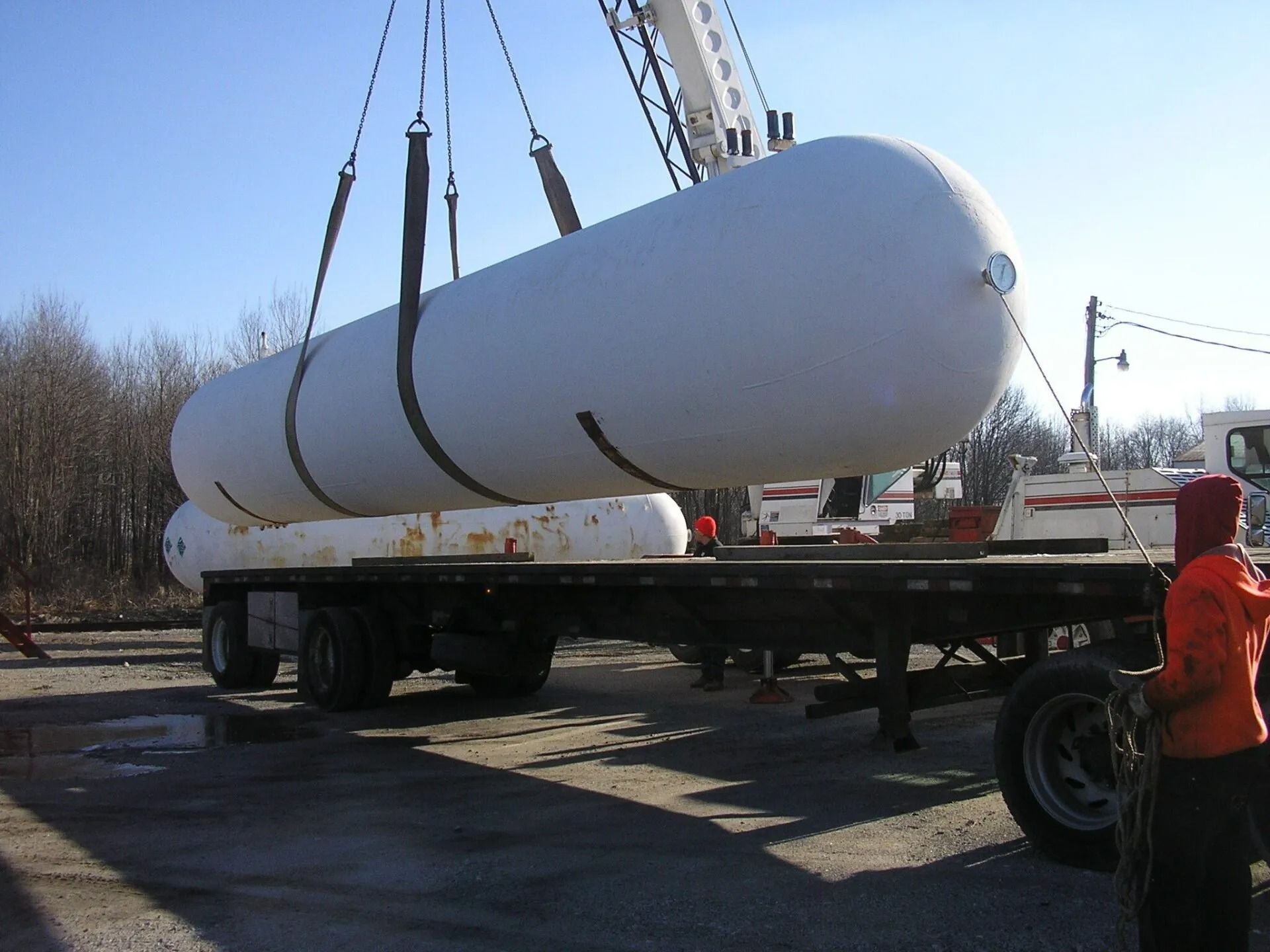 A crane lifts a large white propane tank off a flatbed trailer. A worker in orange is visible.