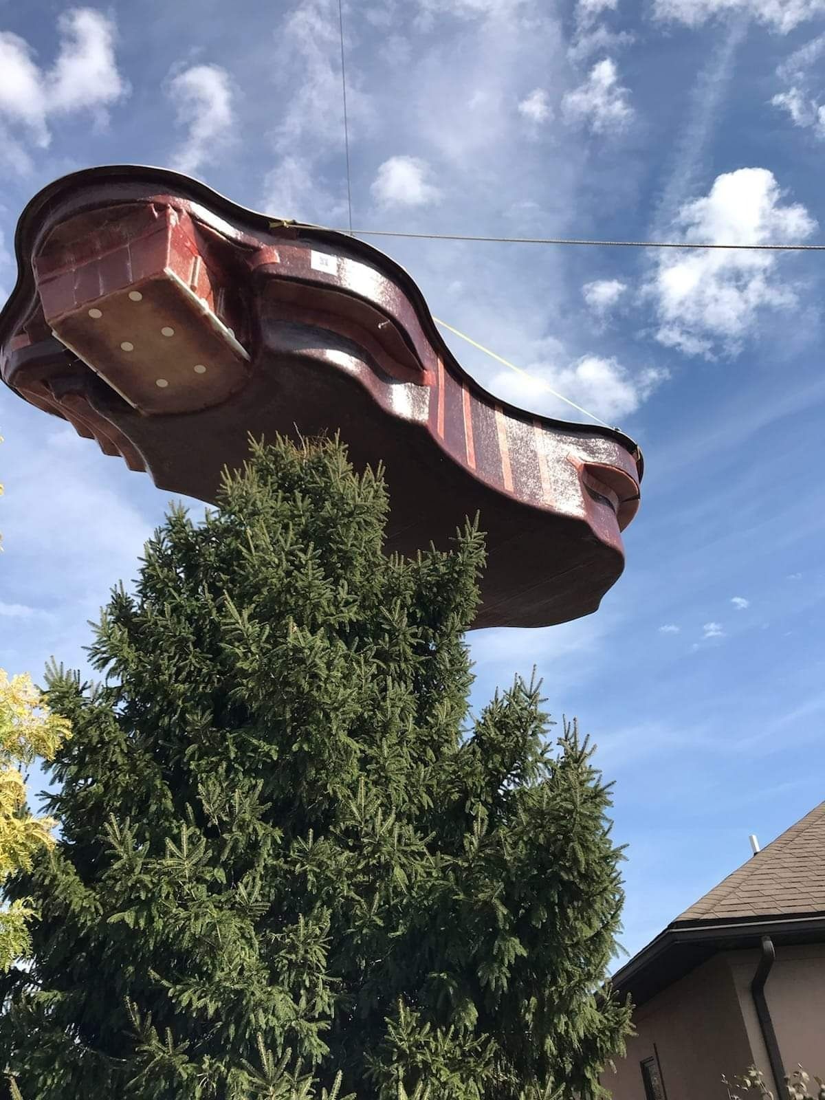 A large, red, heart-shaped hot tub is lifted by cables above a tall evergreen tree and a house, against a cloudy sky.