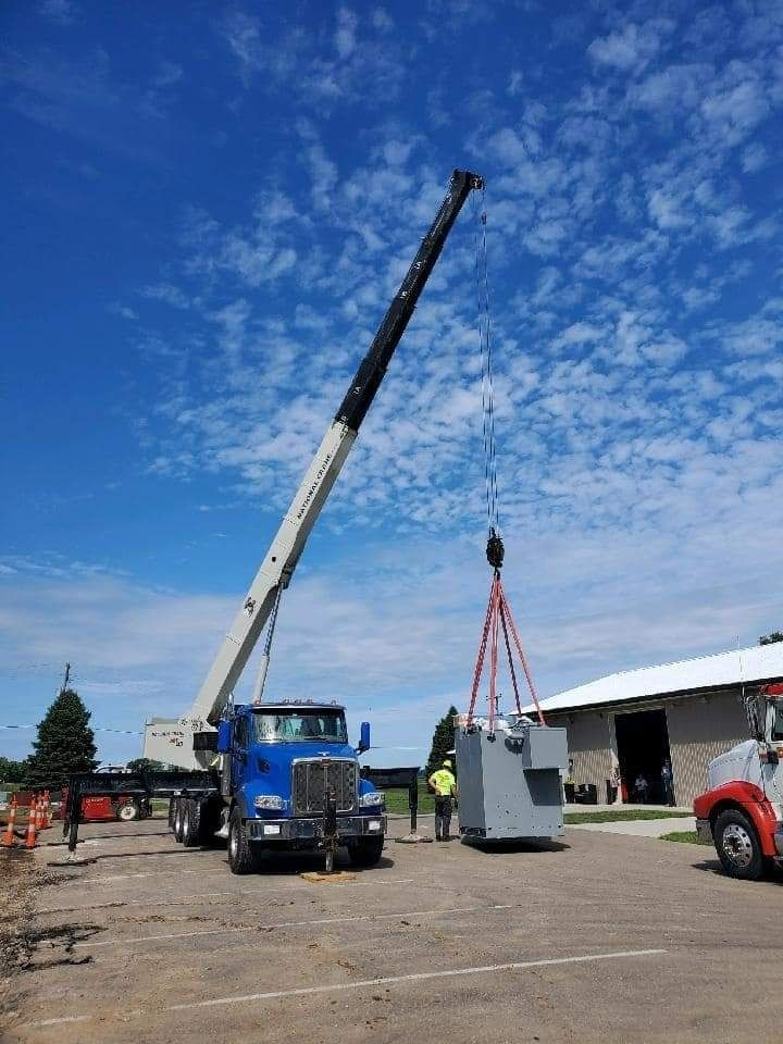 Crane lifting a large gray electrical transformer. A worker stands nearby. Blue truck, white building. Bright blue sky.