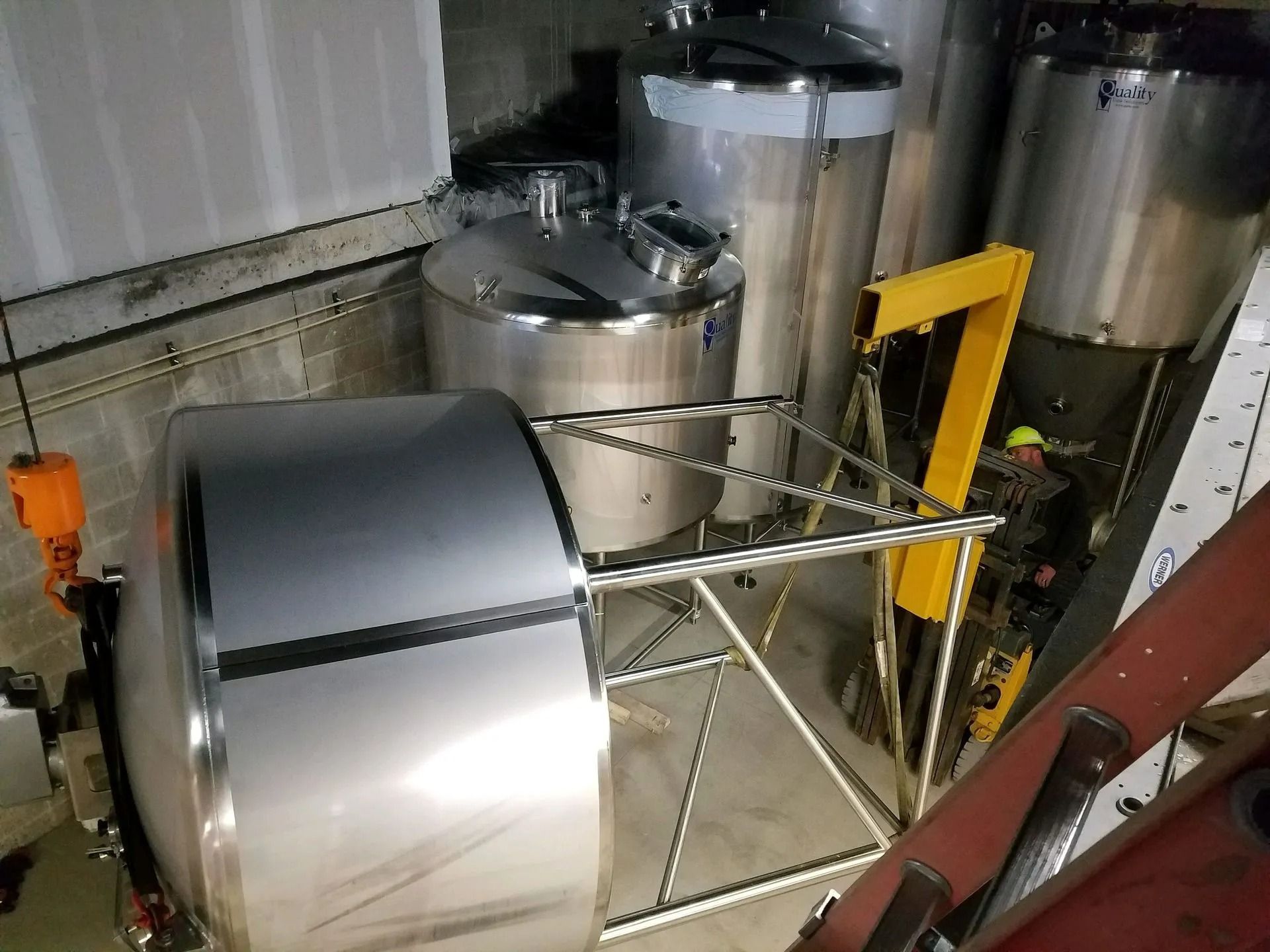 Stainless steel brewing tanks in an industrial setting. Some tanks have lids. Yellow support structure visible.