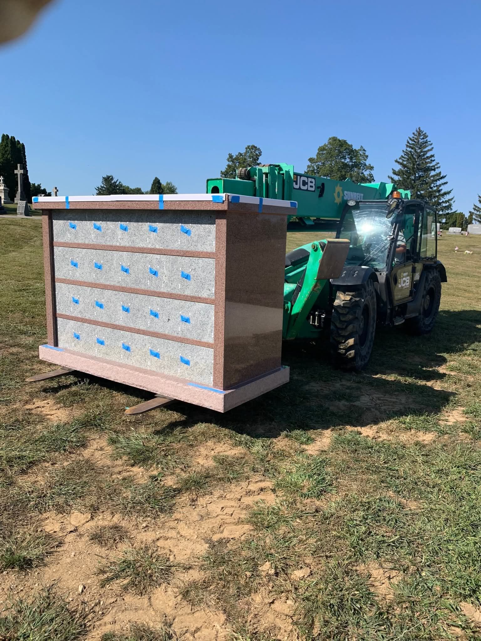 A green forklift moving a large, multi-compartment columbarium in a cemetery on a sunny day.