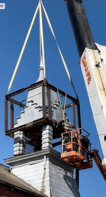 Crane lifting a steeple from a building, worker in a lift basket nearby.