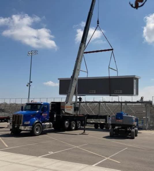 A crane lifting a gray building structure from a blue truck. In a parking lot near a stadium.
