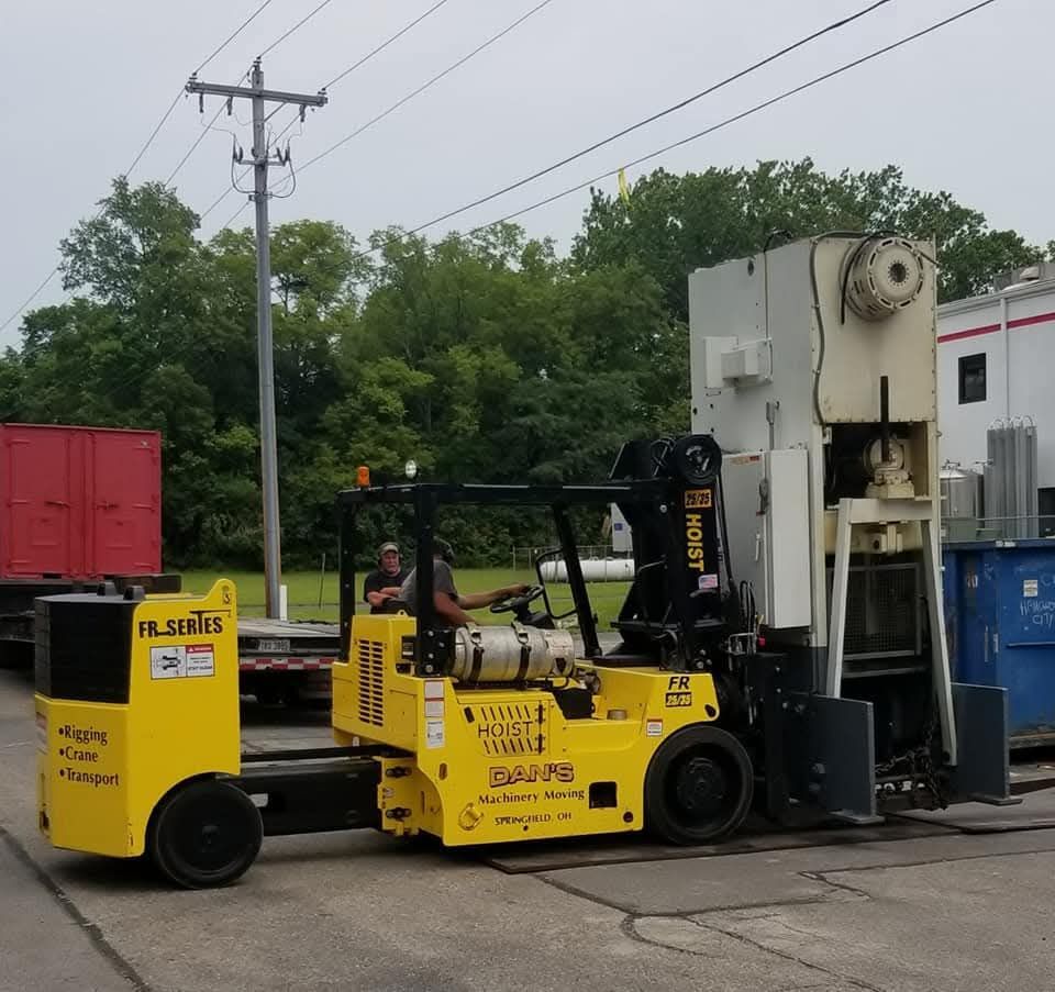 Yellow forklift carrying a large metal industrial machine on pavement; a person is driving.