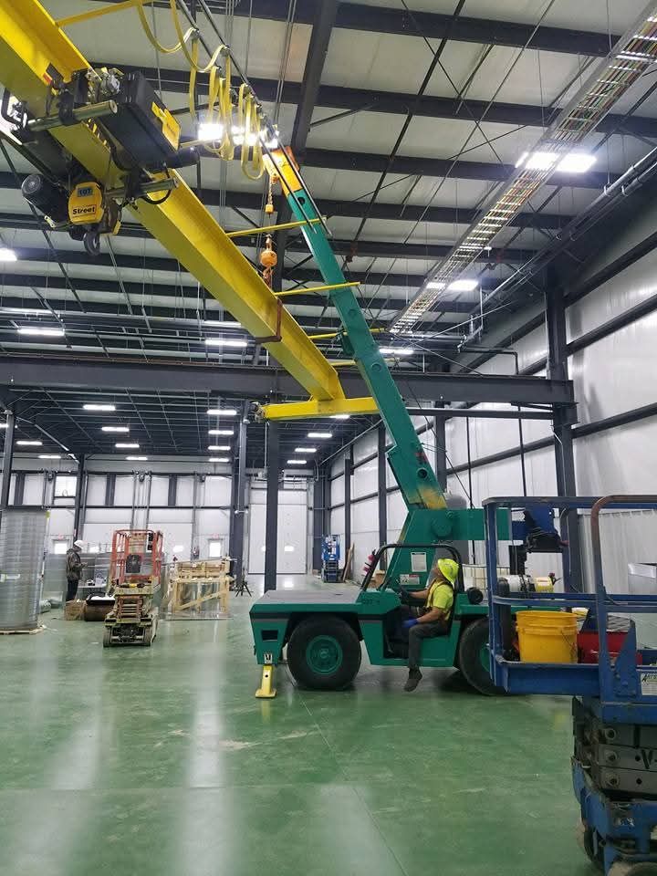 A person operating a green industrial crane. The crane's arm extends up to a yellow overhead beam. An industrial setting.