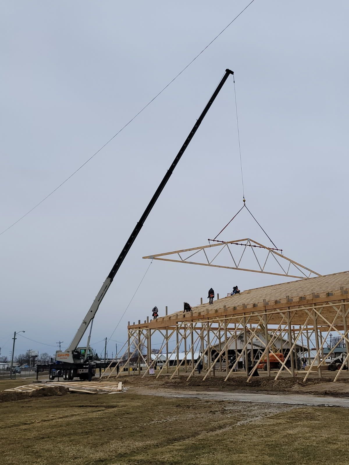 Construction site. Crane lifts roof truss onto wooden building frame. Workers on roof. Cloudy sky.