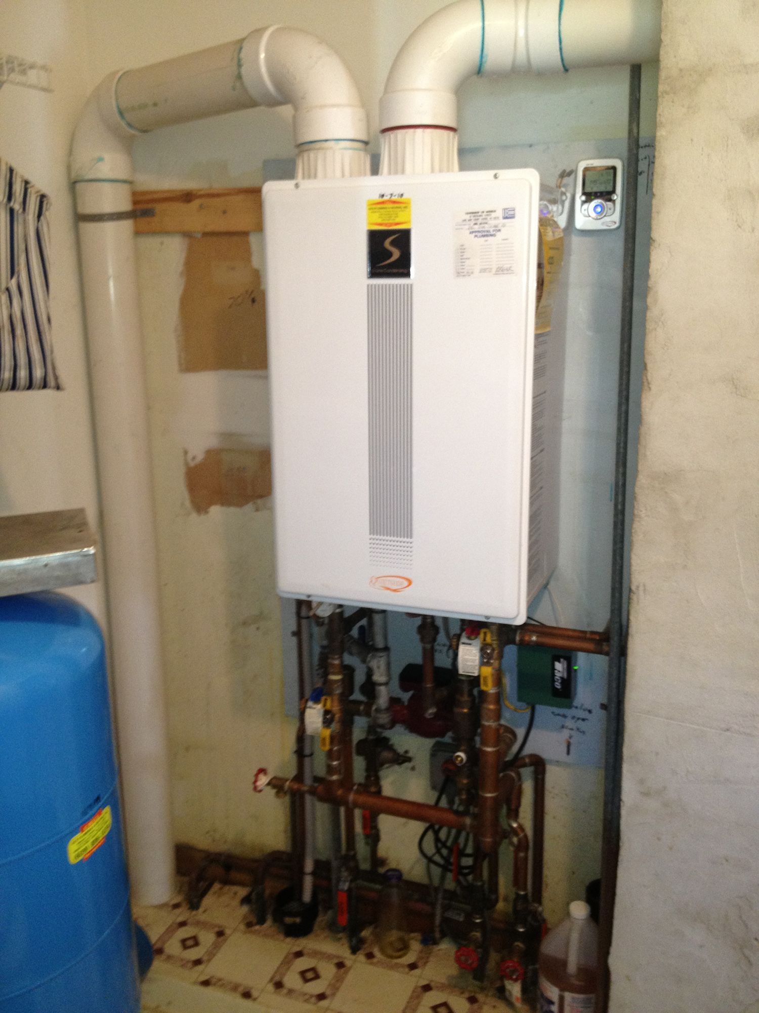 A wall-mounted tankless water heater with PVC intake and exhaust pipes in a utility area next to a blue water tank.