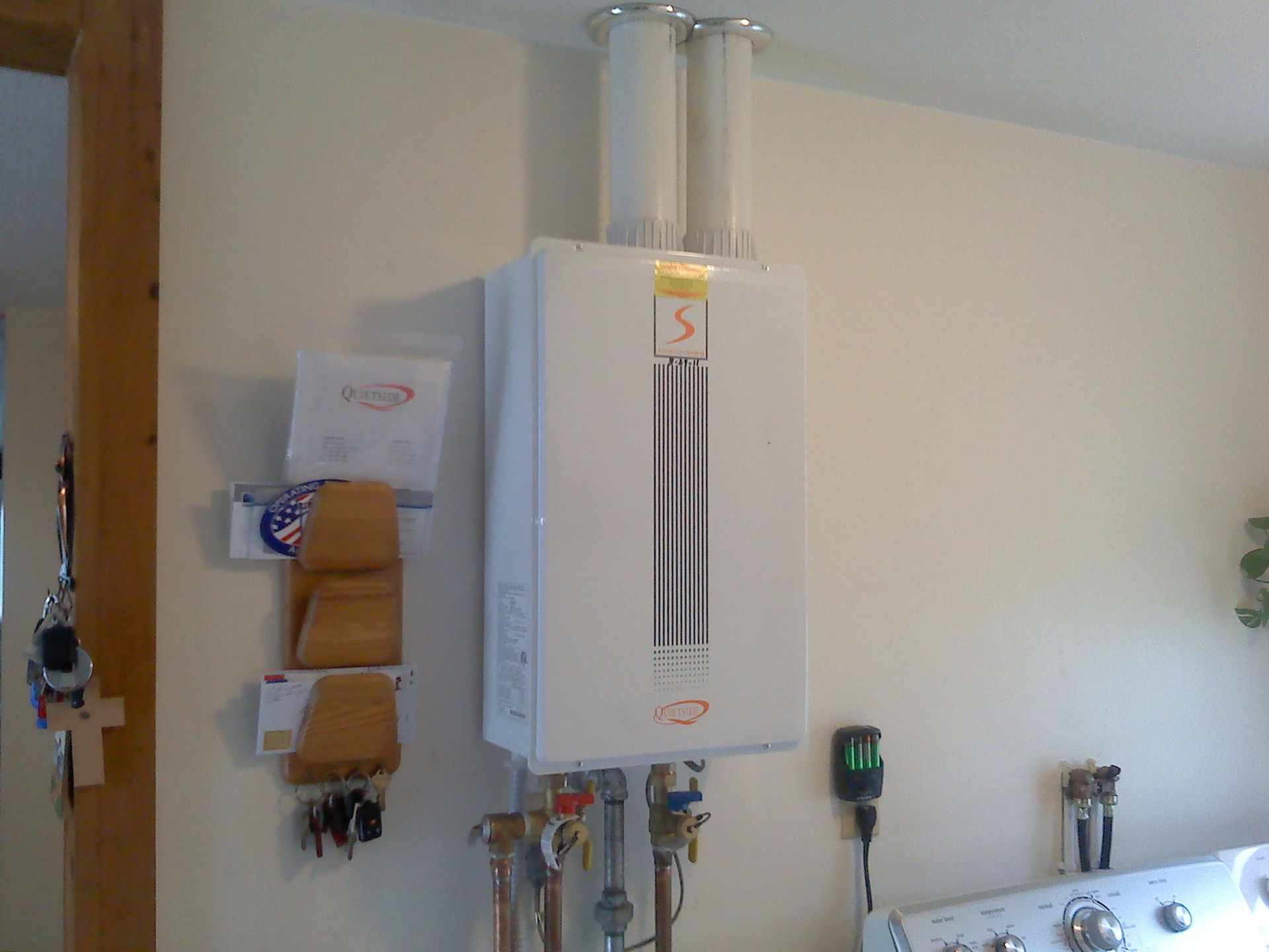 A wall-mounted white Rinnai tankless water heater with two vertical exhaust pipes on a plain wall in a utility room.