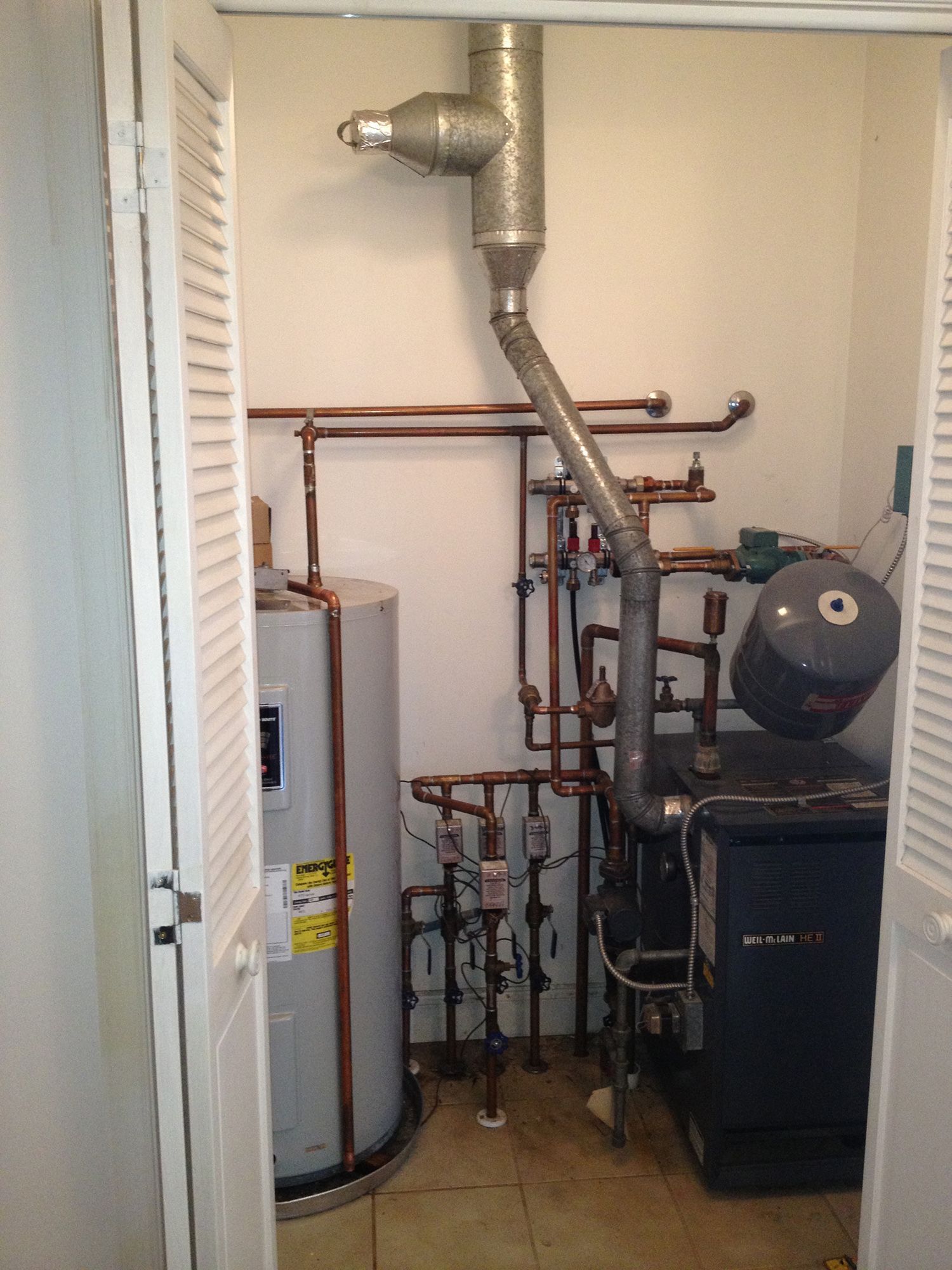 A water heater and a boiler with copper pipes and a metal vent pipe installed in a closet with louvered doors.