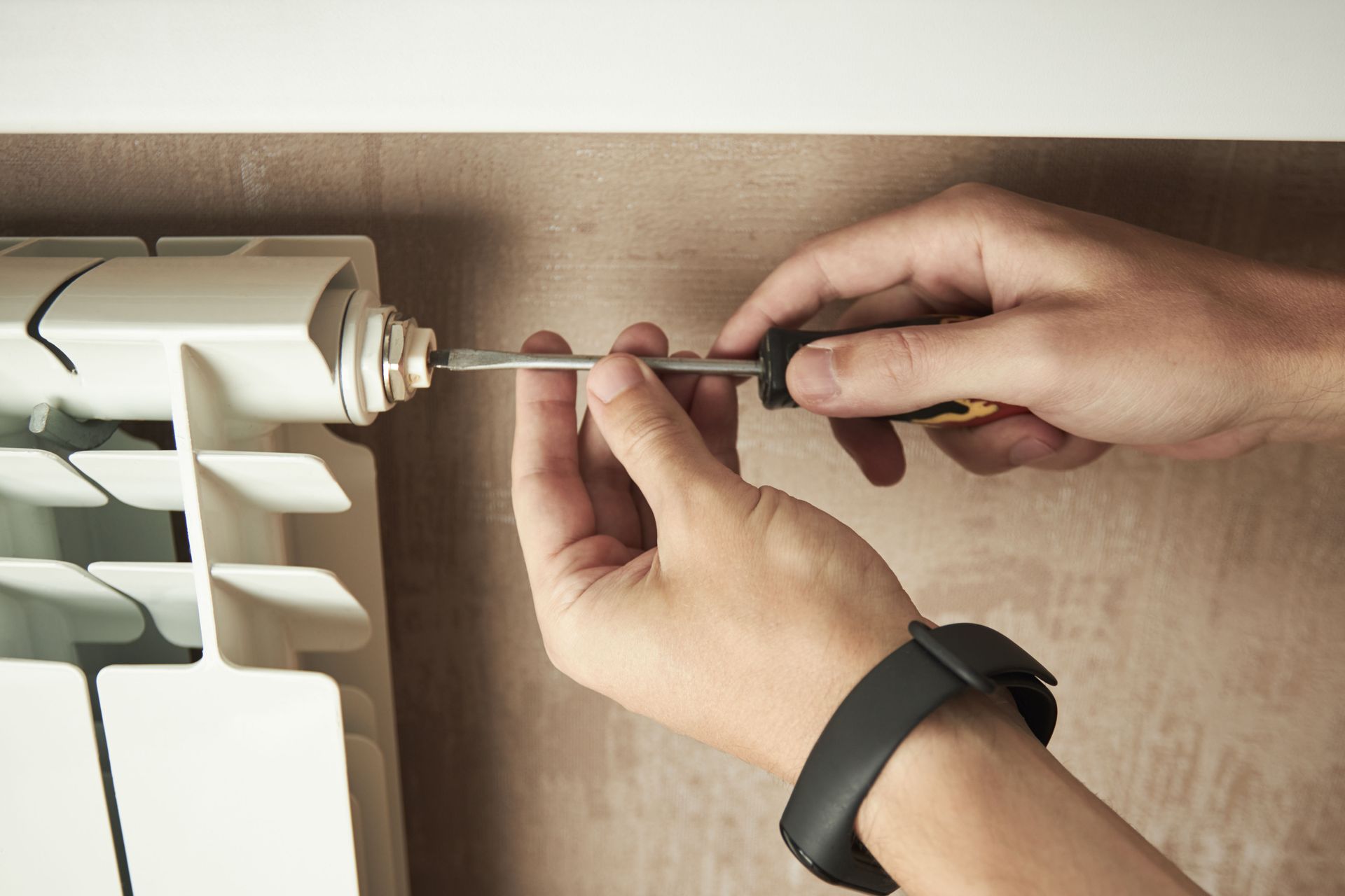 A person uses a screwdriver to tighten a fitting on a white metal radiator mounted against a beige wall.