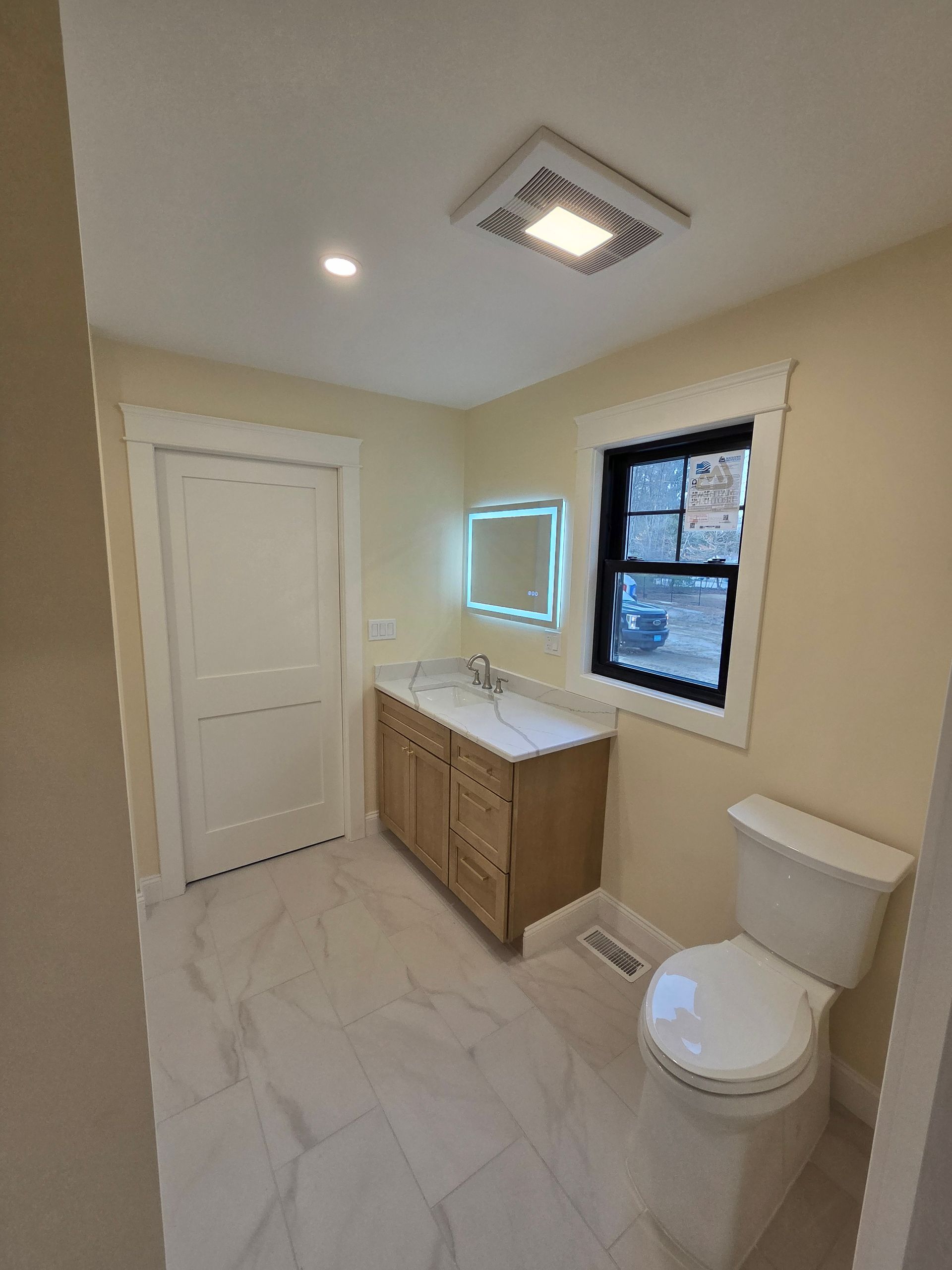 A bathroom with a toilet, sink, mirror, and window.