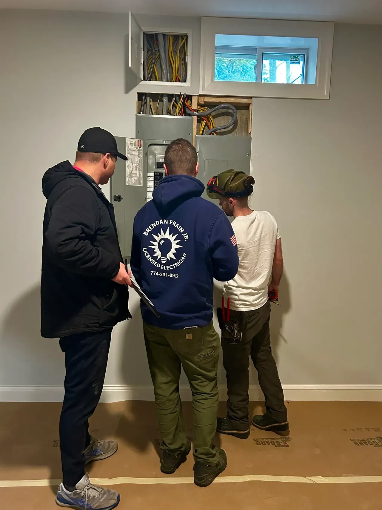 Three men are standing in a room looking at a electrical box.
