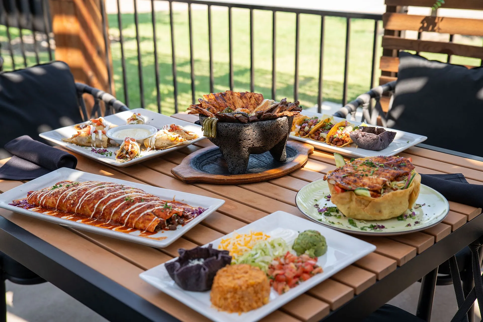 Mexican food spread on a wooden table outdoors: enchiladas, tacos, and other dishes.
