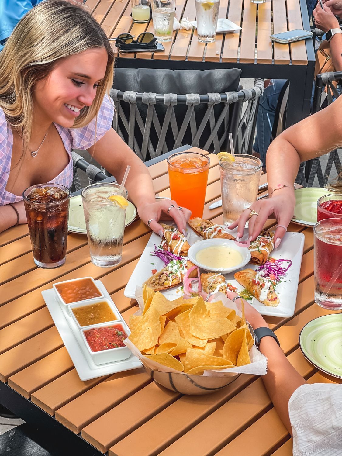 People at an outdoor table sharing food and drinks. Woman smiling, reaching for the food. Chips, salsa, and cocktails on the table.