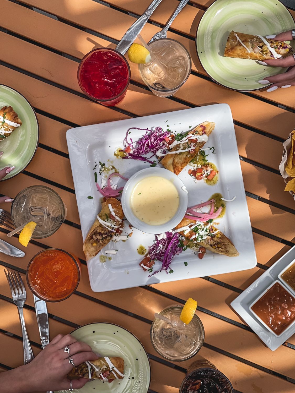 Overhead view of a table with food, drinks, and hands. Plates of food surround a central plate with a dipping sauce.