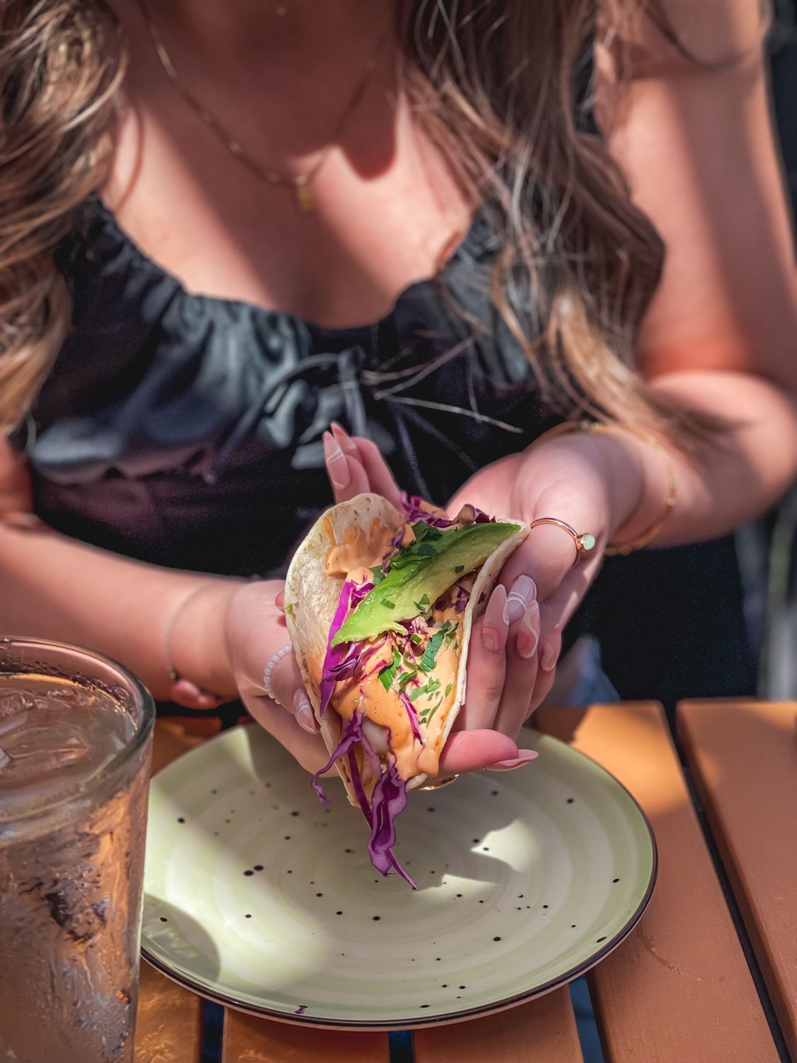 Person holding a taco with avocado and purple cabbage; on a plate with a drink.