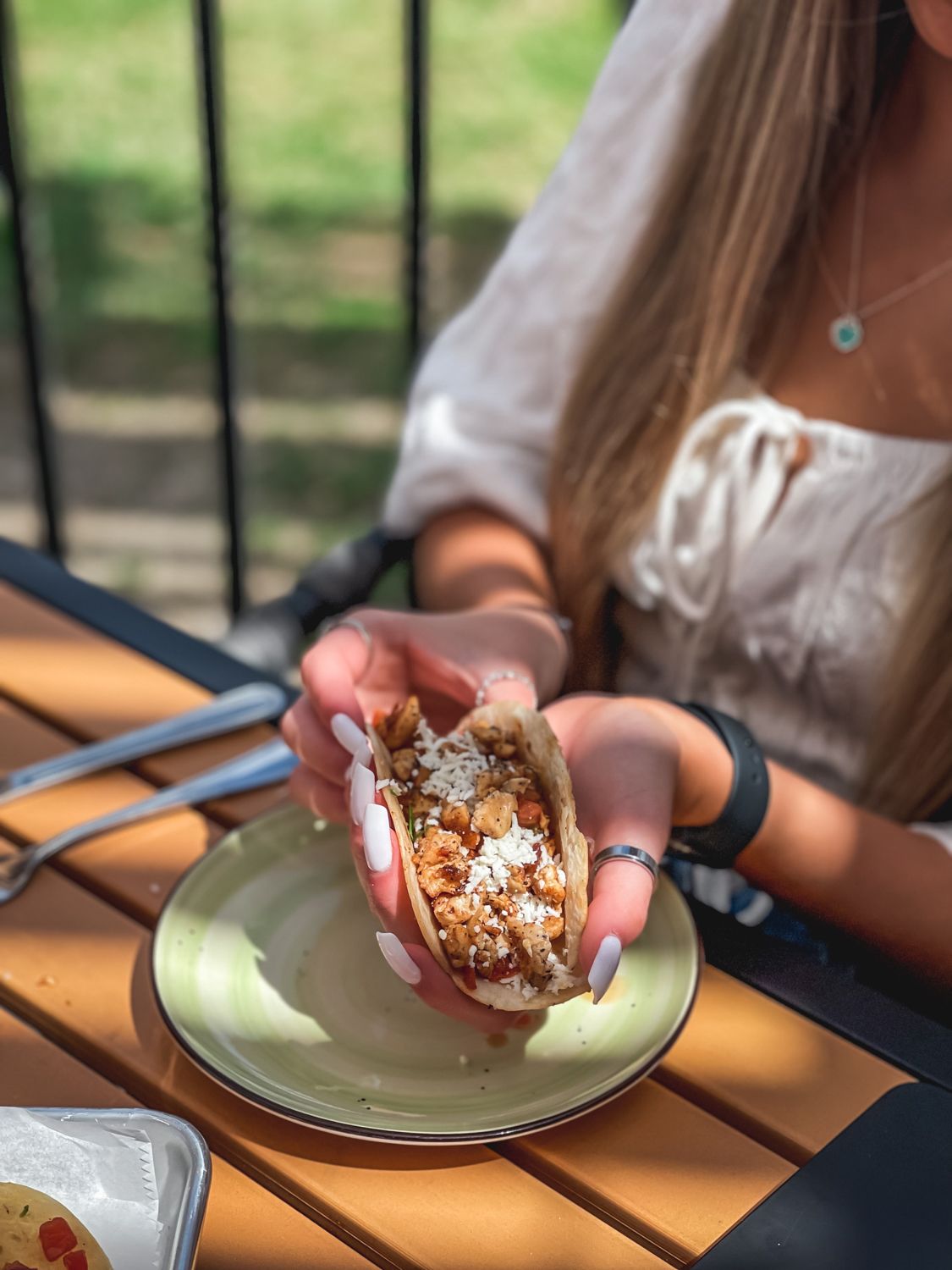Woman holding a taco, with a plate and utensils on a wooden table outdoors.