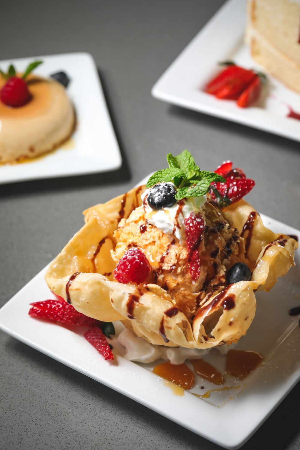 Fried ice cream dessert in a fried tortilla bowl with fruit, cream, and chocolate drizzle; other desserts in background.