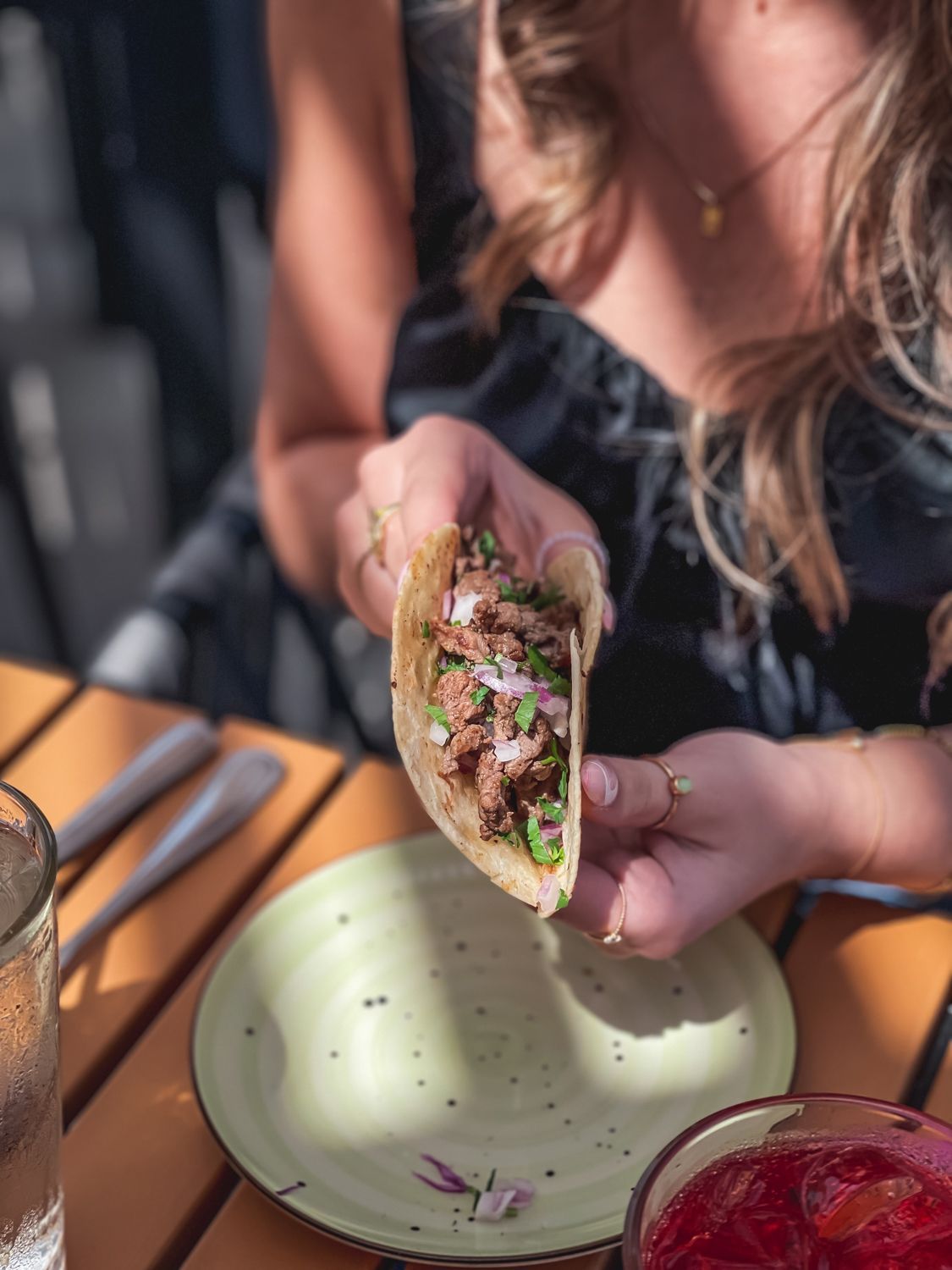 Person holding a taco at an outdoor table. Taco contains meat, onions, and cilantro. Green plate and glass visible.
