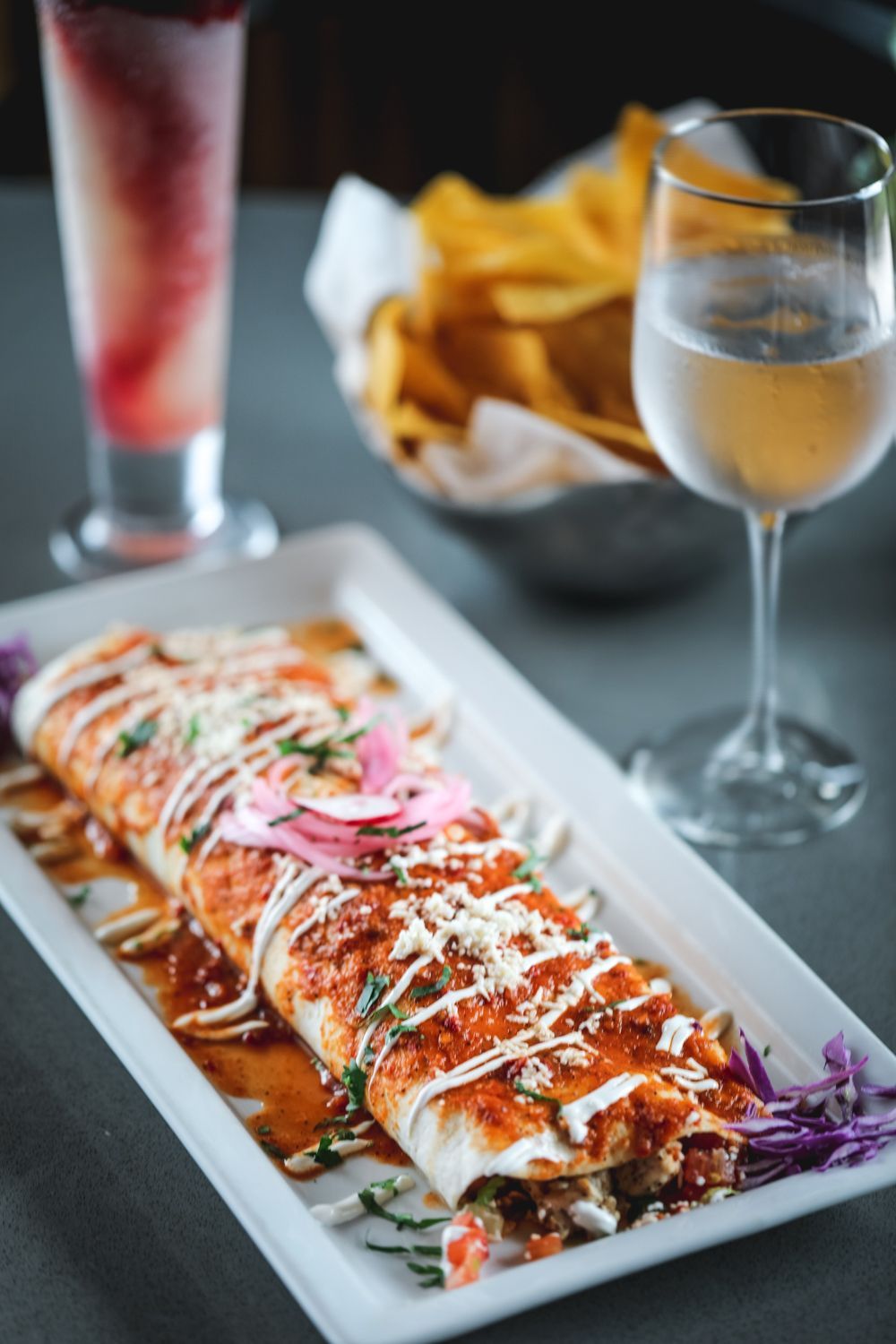 Burrito with sauce, cheese, and onions on a white plate, served with chips, a cocktail, and white wine.