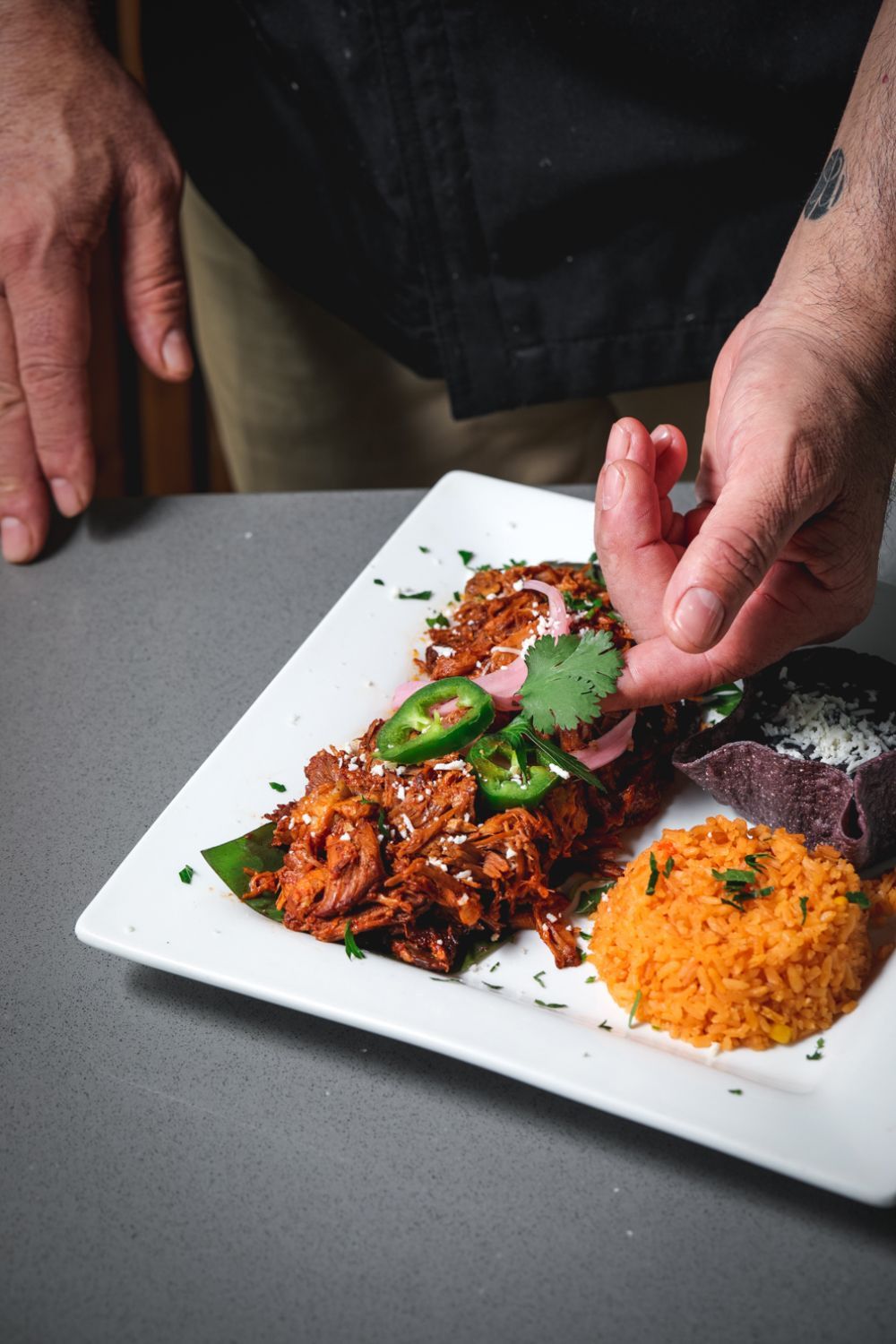 Chef garnishes a plate of shredded meat with jalapeños, cilantro, rice, and purple tortillas.