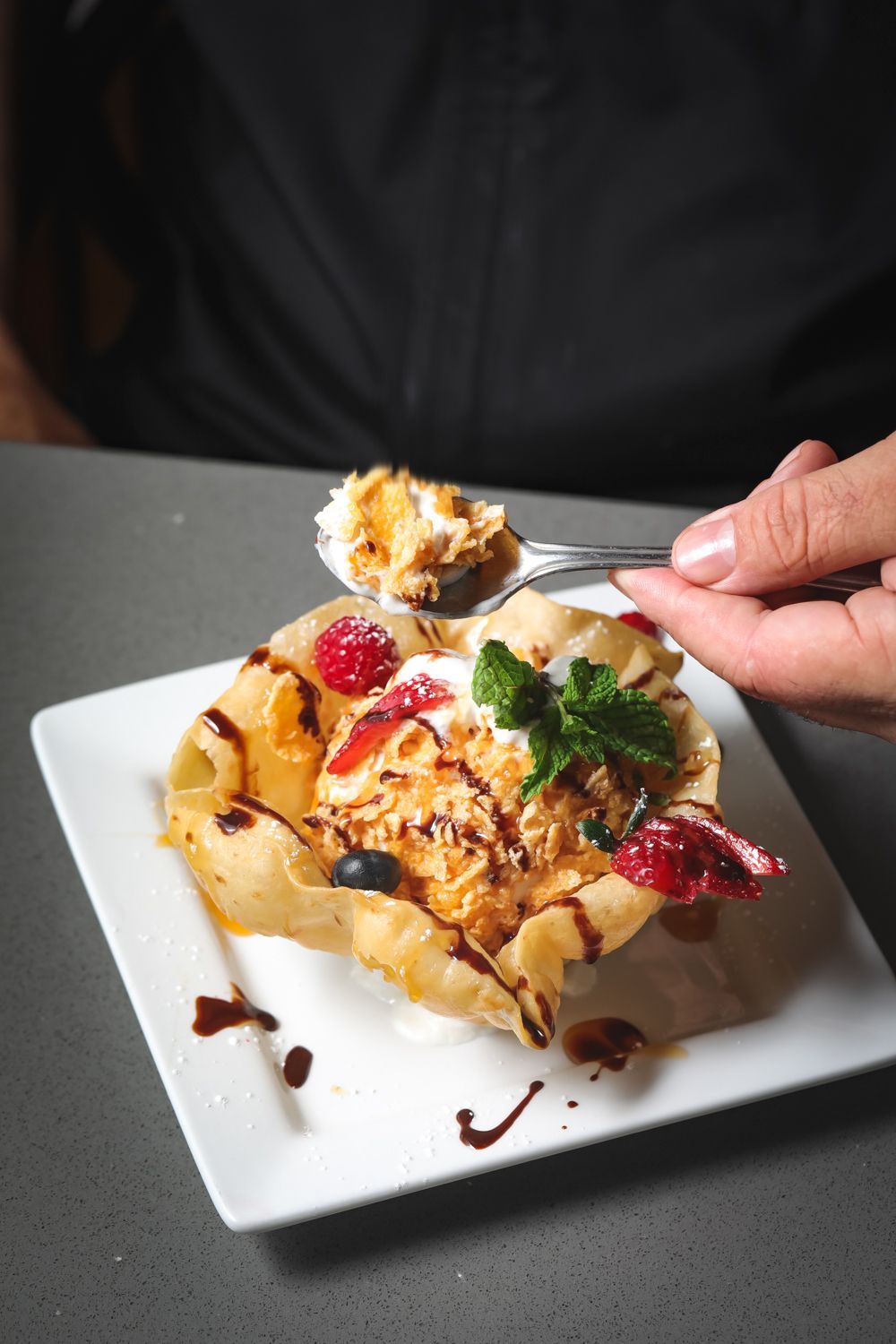 Dessert in a fried tortilla bowl with ice cream, fruit, whipped cream, and sauce. Someone is eating it.