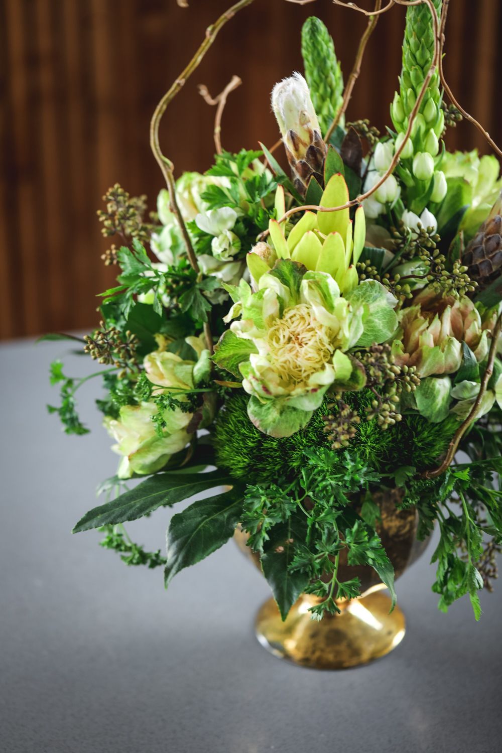 Green and white floral arrangement in a gold-rimmed pedestal vase, against a wooden background.