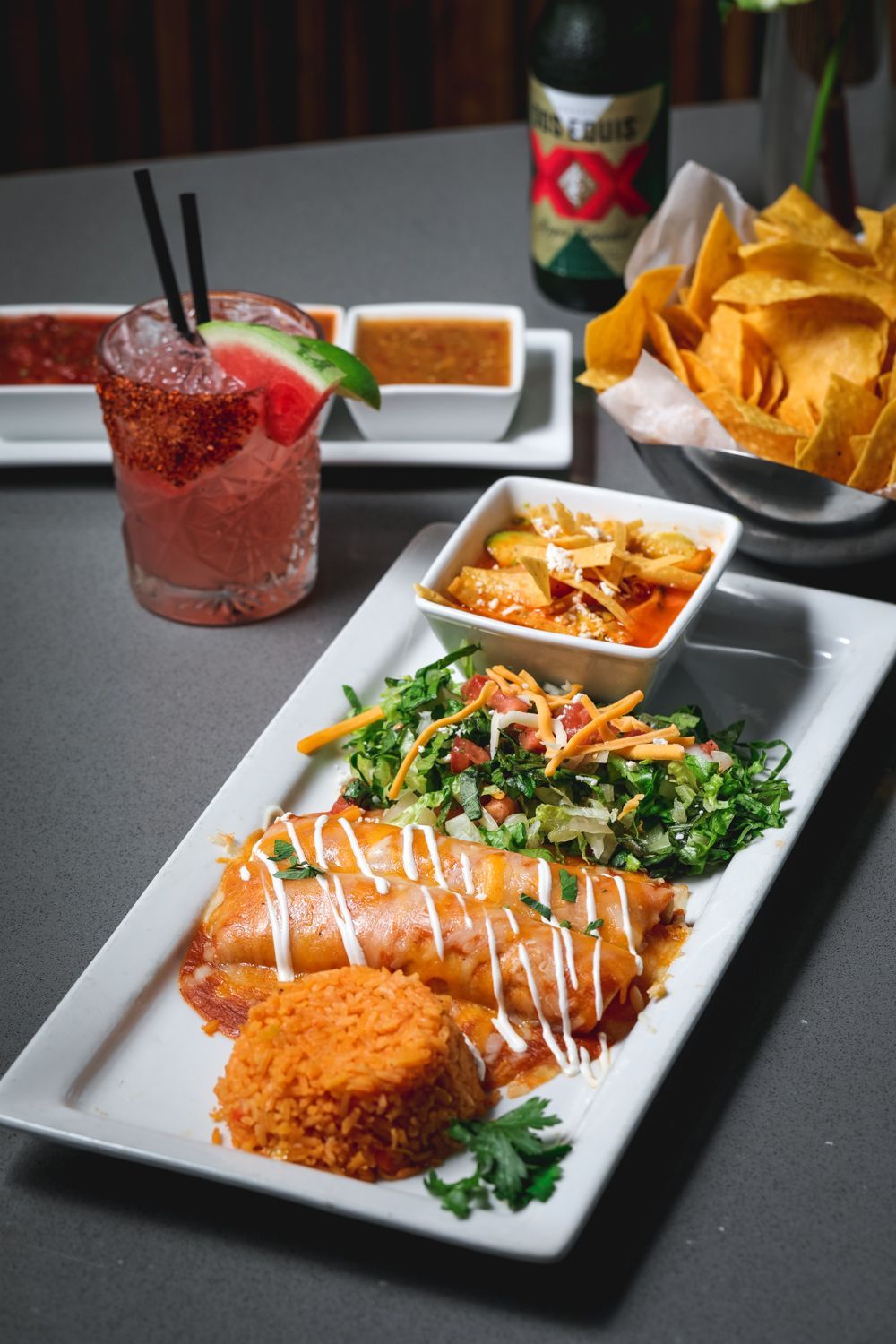 Enchiladas with rice and salad on a white plate, alongside chips, salsa, and a watermelon cocktail.