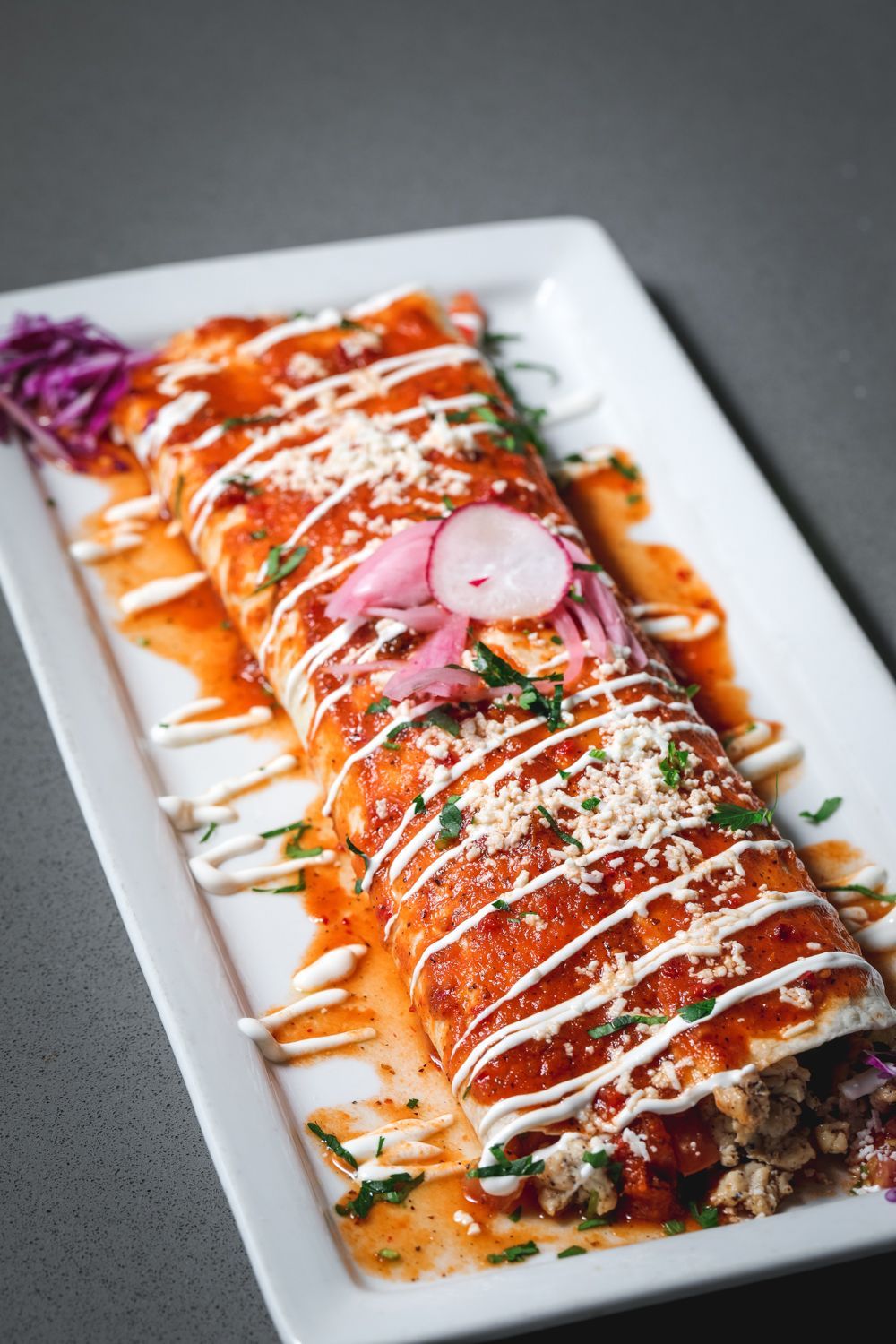 Enchilada-style burrito on a rectangular white plate, covered in sauce, crema, cheese, and pickled toppings.